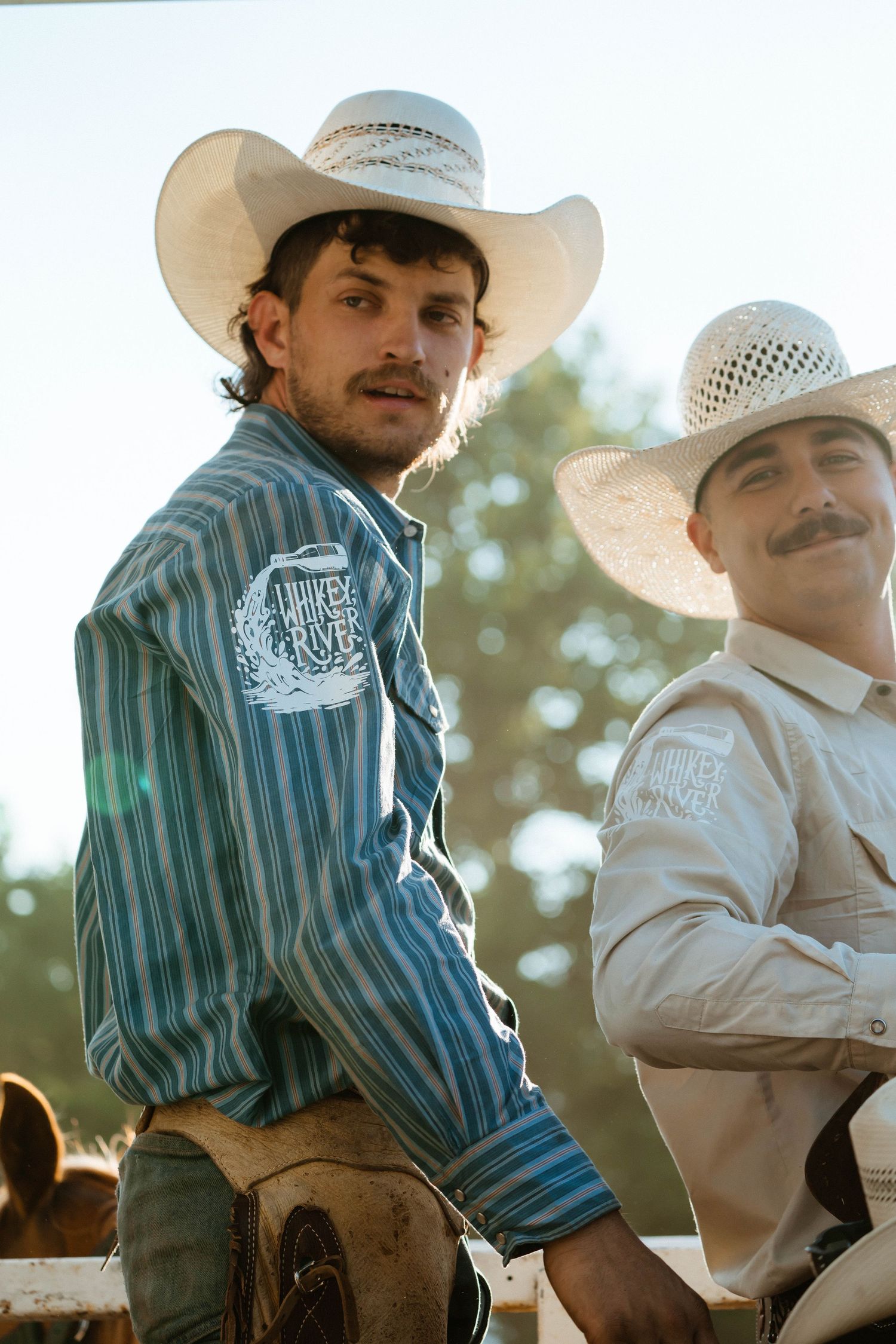 Cowboys in western attire sitting on fence at rodeo grounds during golden hour.