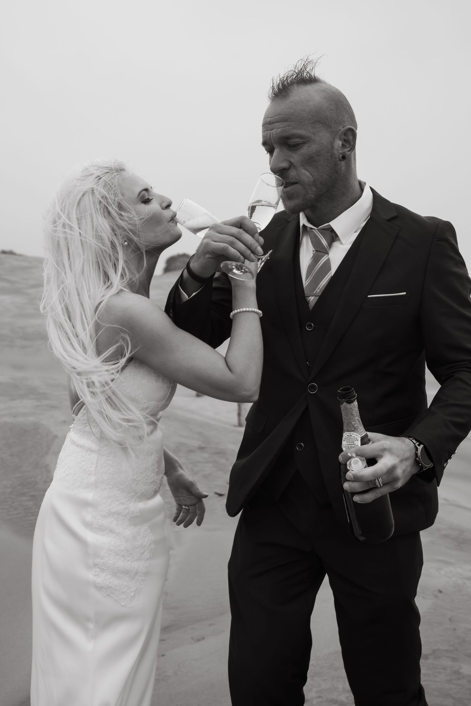 A couple in wedding attire shares a playful moment on a cloudy beach.