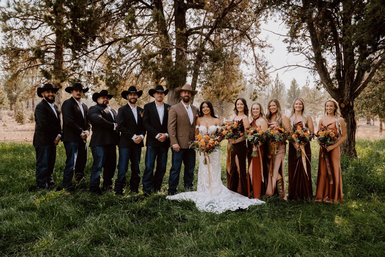 Wedding party in earth-toned attire posing together under tall trees during an outdoor ceremony.