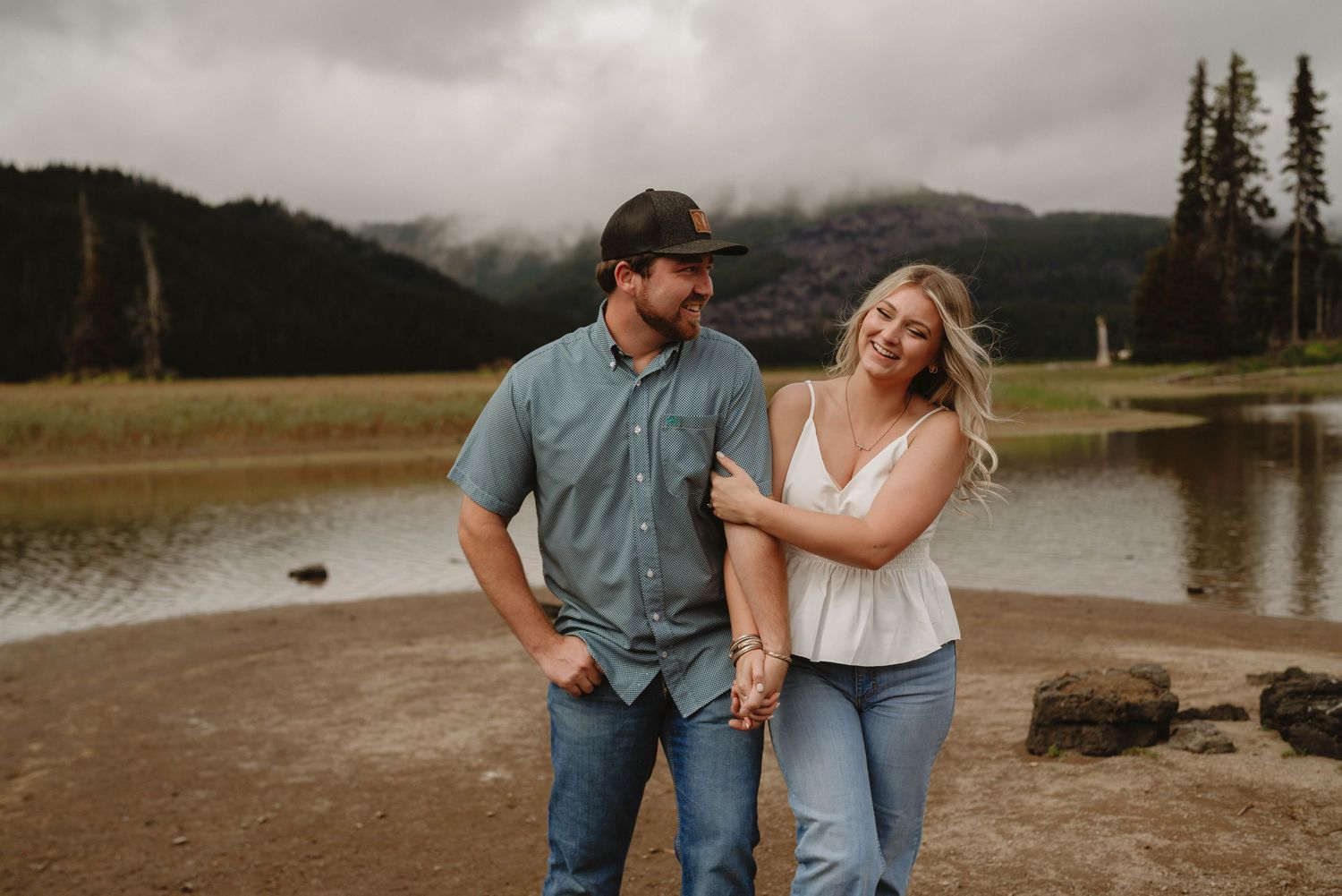 Couple walking together near a scenic mountain lake with evergreen trees in the background.