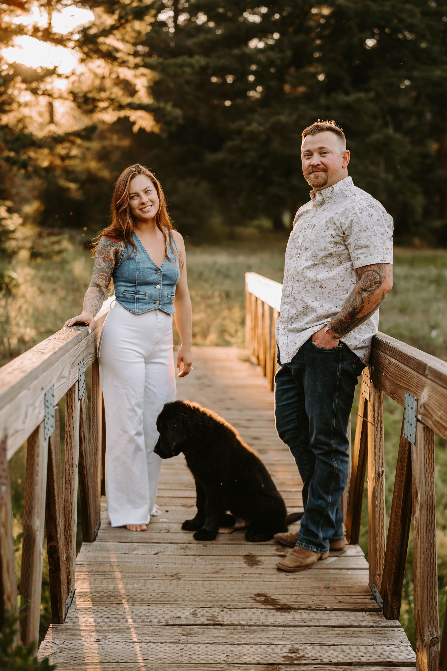 Couple standing on wooden boardwalk with black dog at sunset among pine trees.