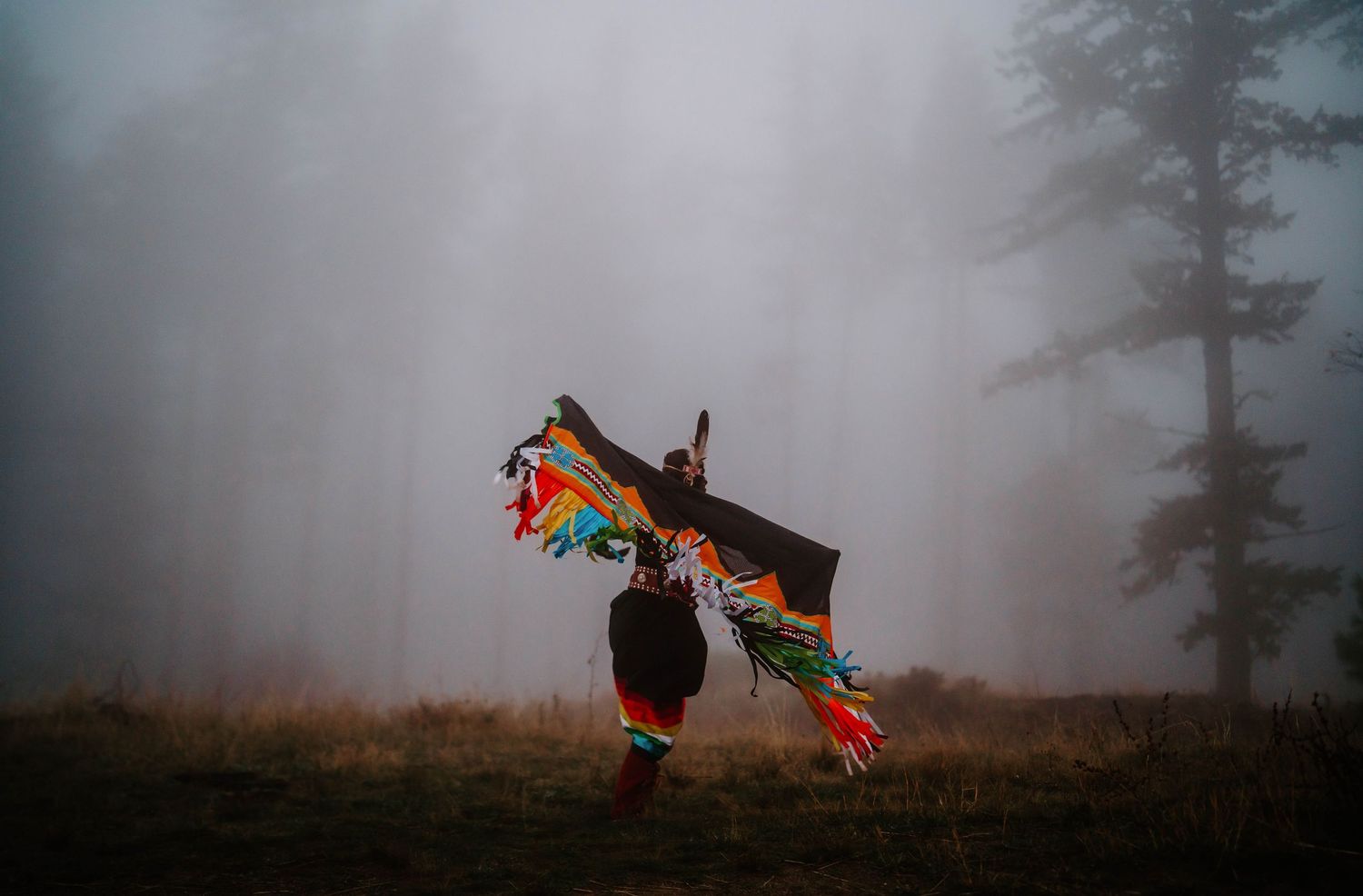 Colorful traditional regalia and fringe details shown in moody fog against a natural landscape.