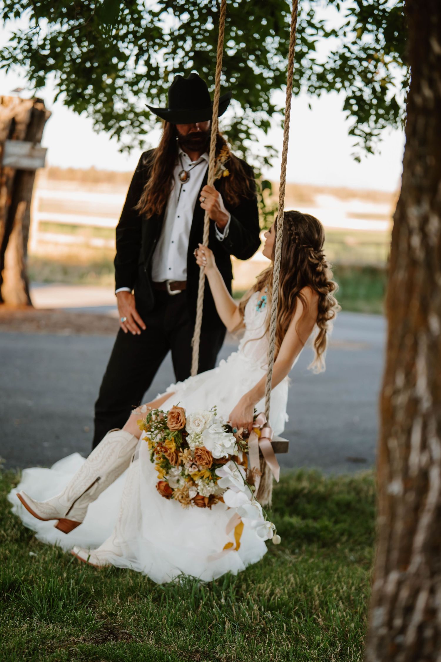 A couple sits on a tree swing in formal attire holding autumn-colored flowers.