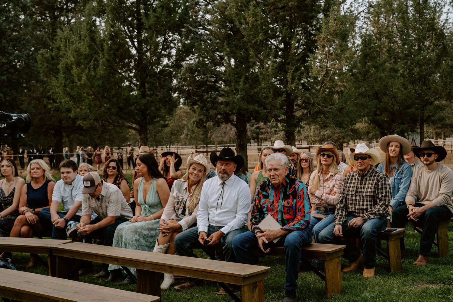 Wide shot of wedding guests seated on wooden benches at an outdoor ceremony venue.