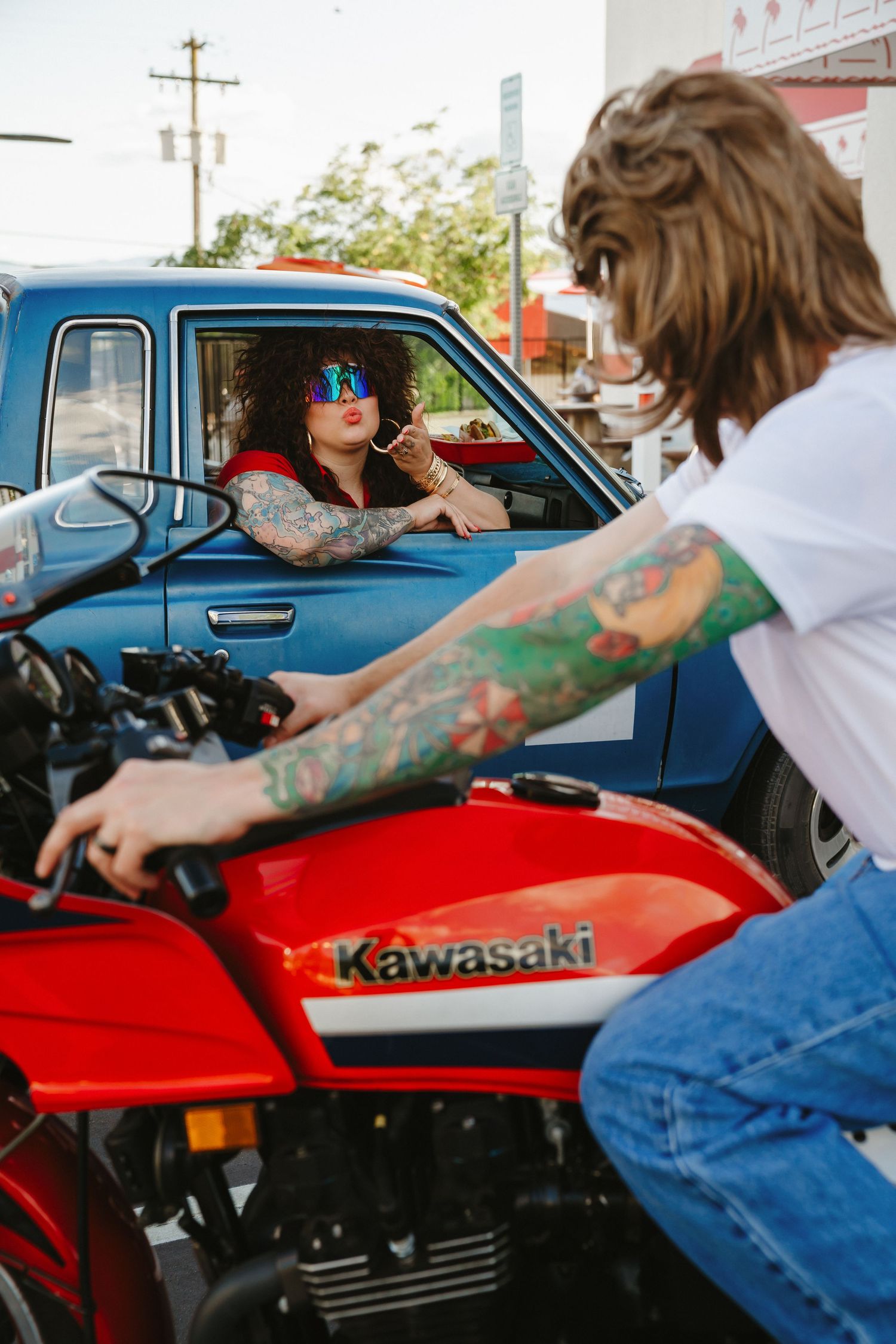 Red Kawasaki motorcycle with tattooed rider in denim jacket outside a vintage car.