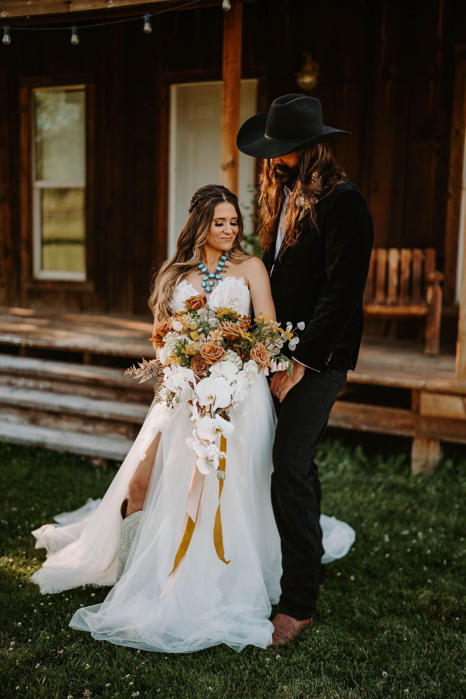 A wedding couple embraces on grass with a stunning floral bouquet featuring white orchids and peach roses.
