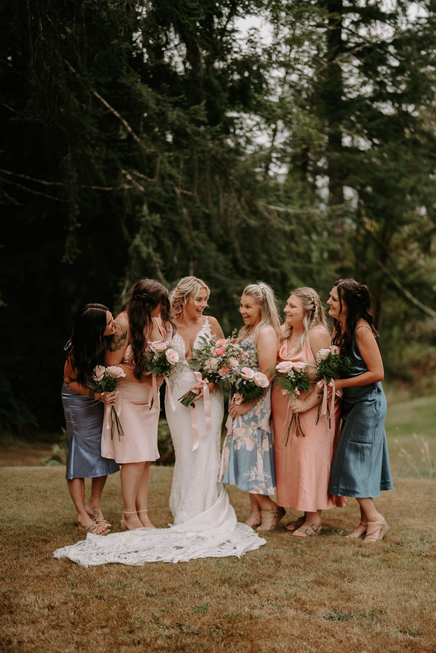 Bridesmaids in pastel dresses gather around the bride on grassy outdoor setting.