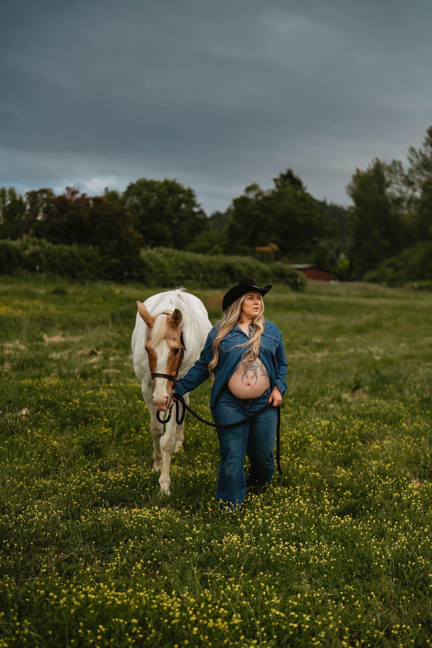 Denim-clad figure poses with white horse in field of yellow wildflowers.