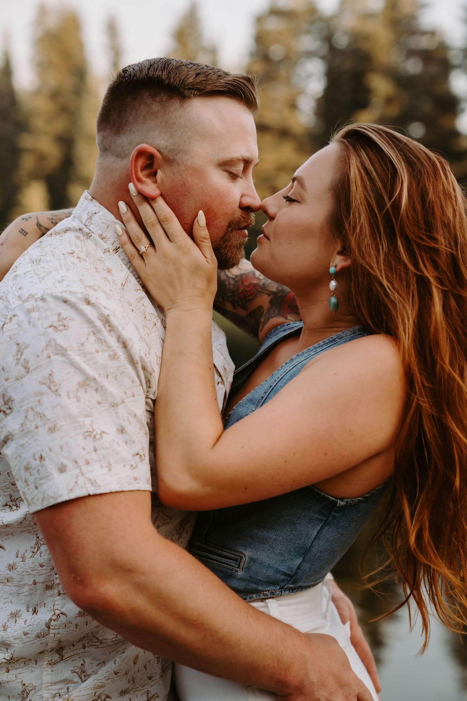 A couple embraces passionately by a lake with pine trees reflected in the water.