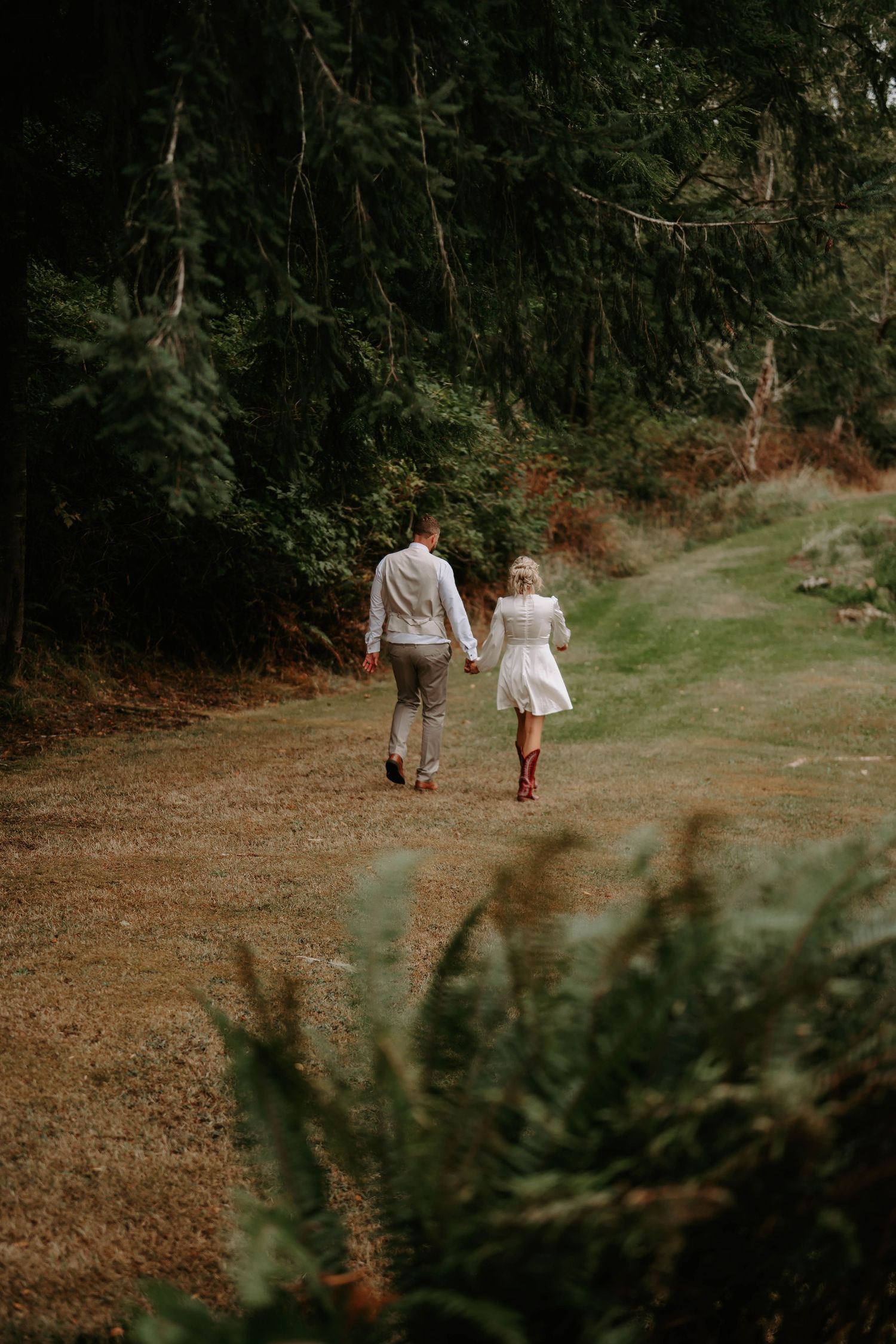A couple walks along a forest path wearing casual attire and rain boots.