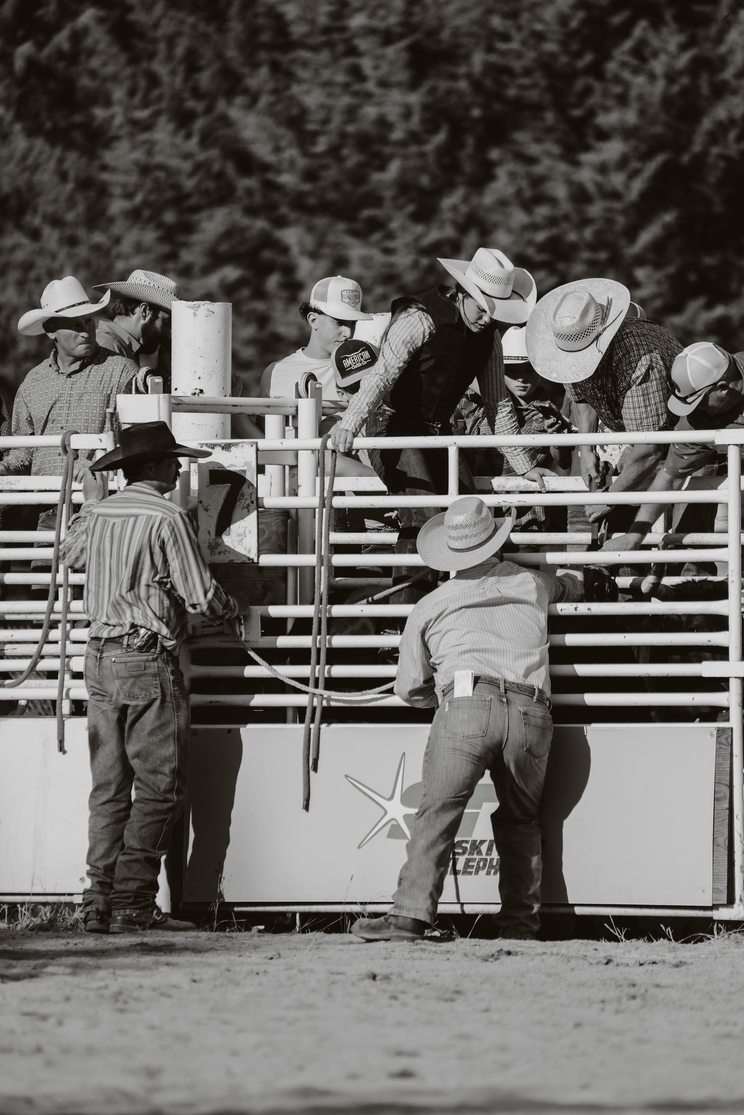 Spectators in cowboy hats lean against white arena fence watching rodeo event.