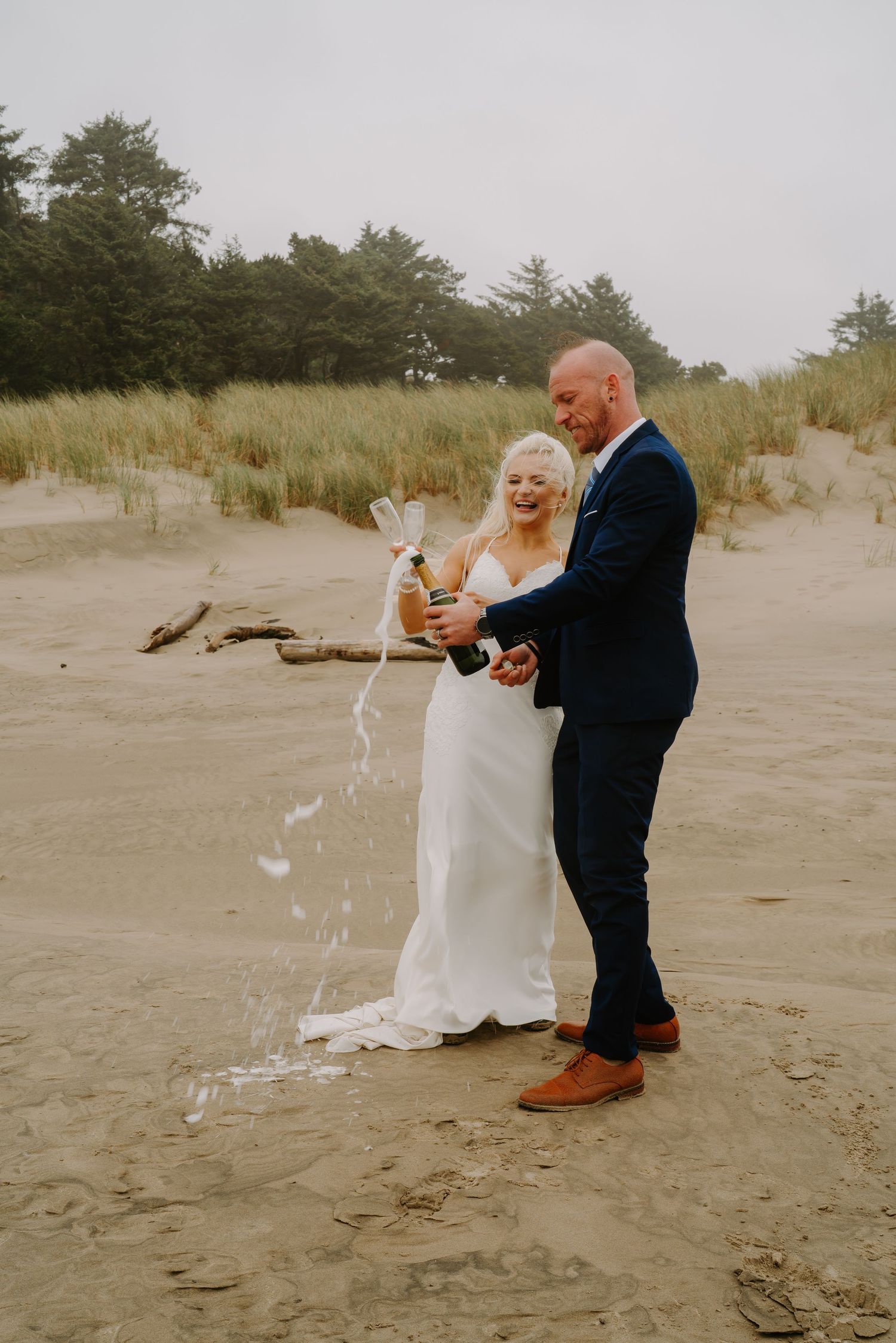 A couple pops champagne while celebrating on a sandy beach with dunes in the background.