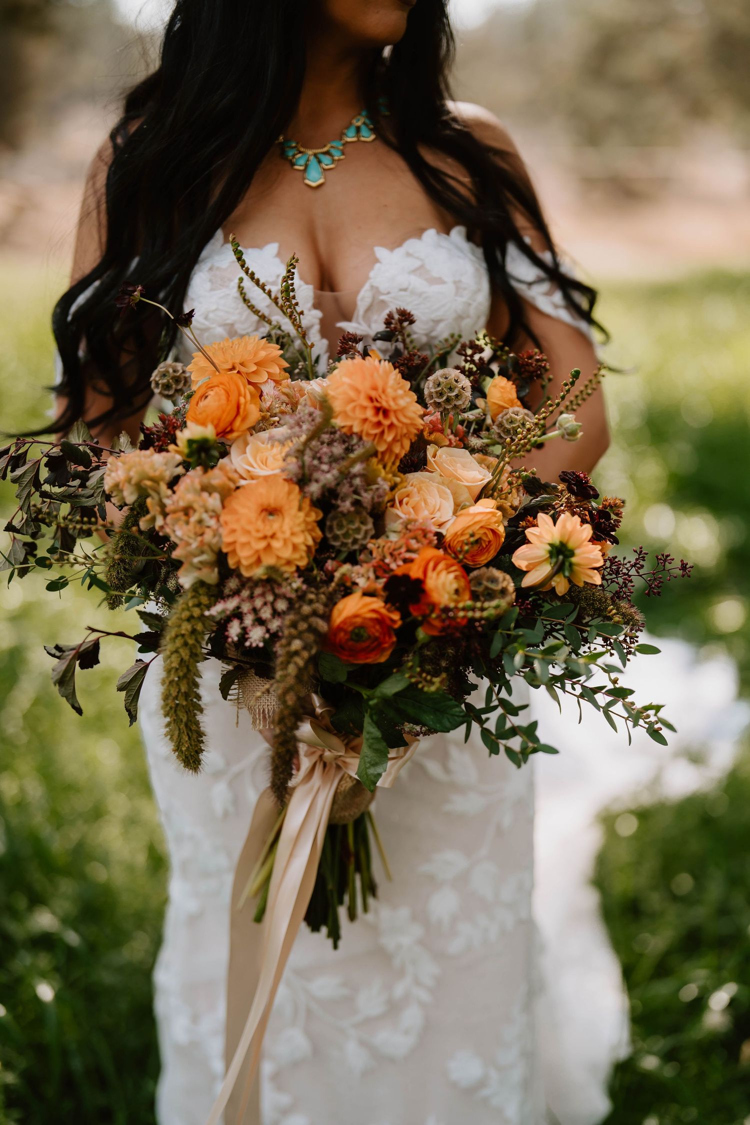 Stunning autumn-colored bridal bouquet with orange dahlias, roses and greenery against a white lace dress.
