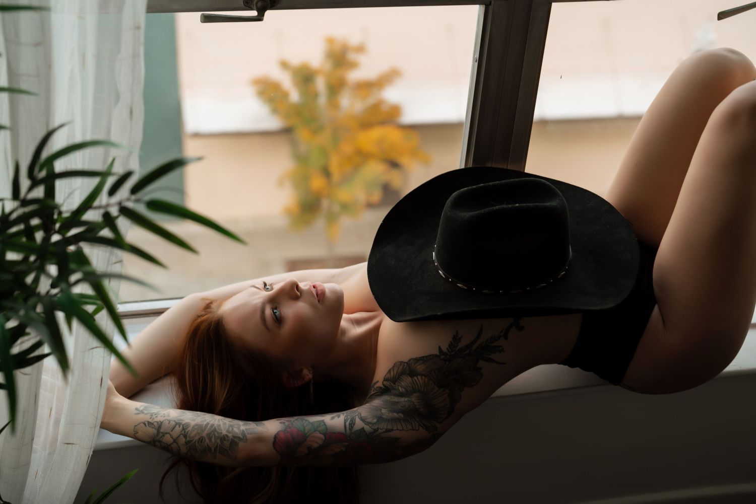Person in black lingerie relaxes by window with indoor plant and autumn tree visible outside.
