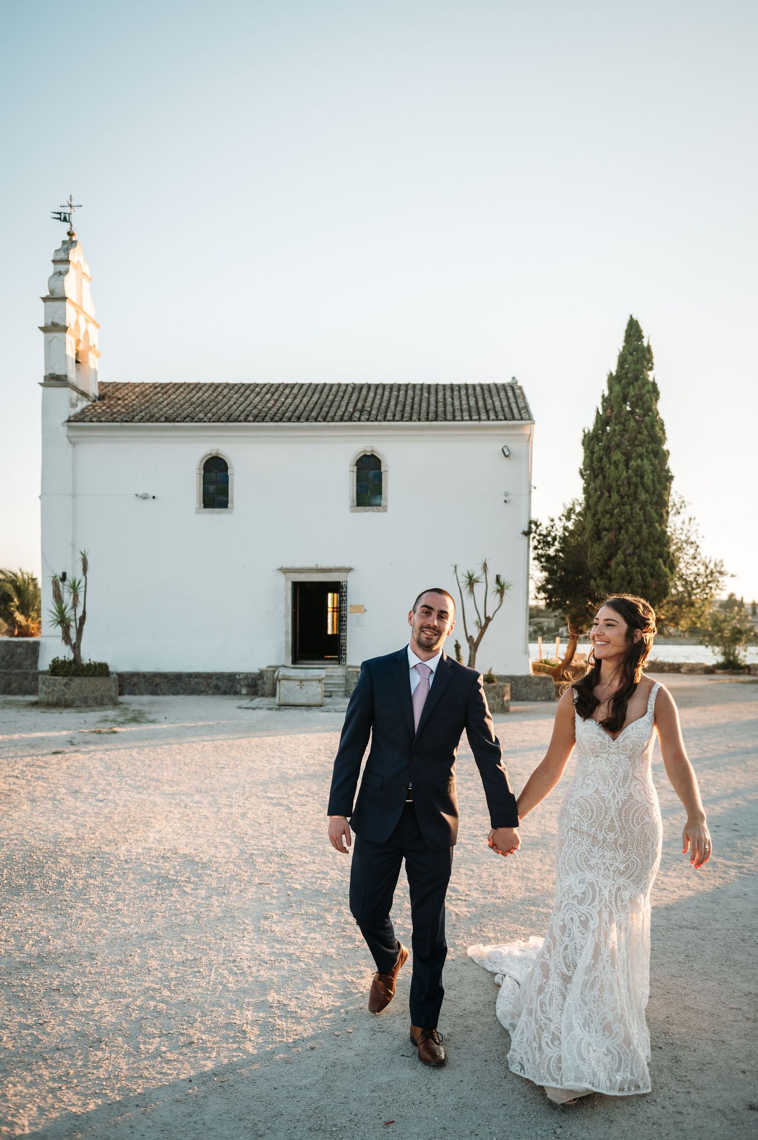 A wedding couple walks together in front of a charming white Mediterranean church.
