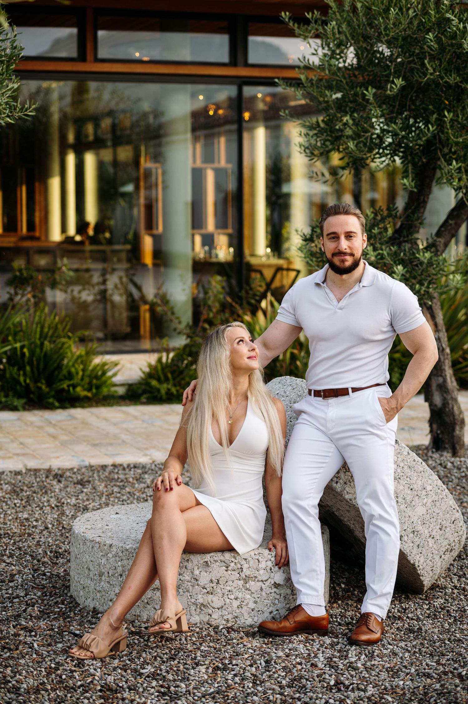 A couple in matching white outfits poses casually on a rocky beach setting.