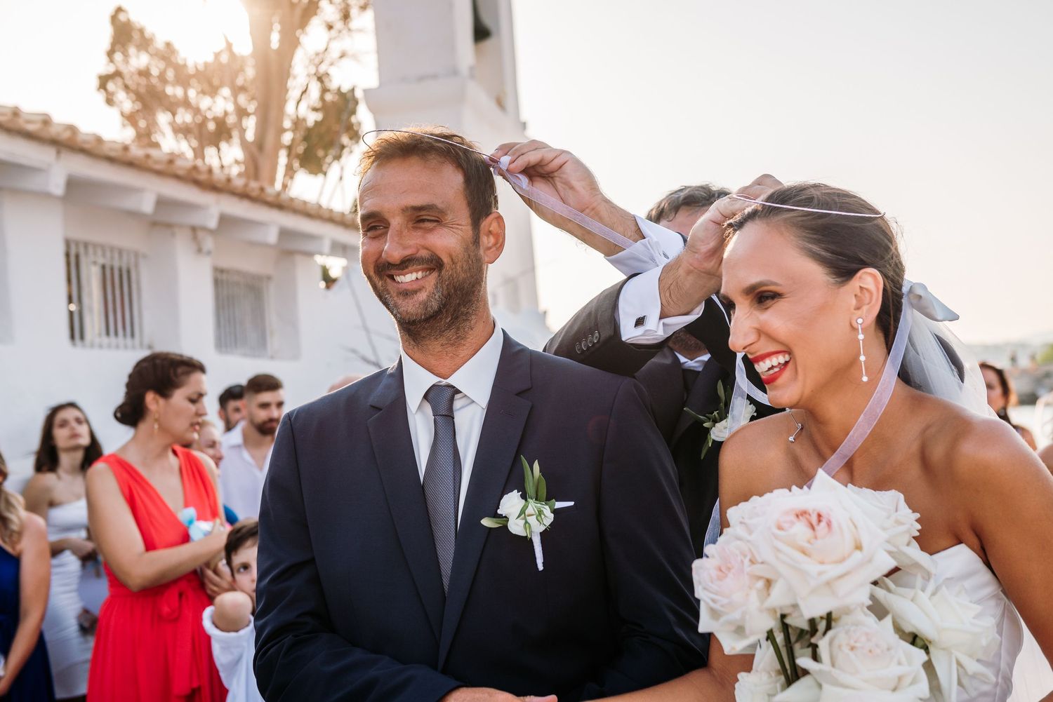 Wedding party celebrating outdoors at sunset with white Spanish-style building in background.