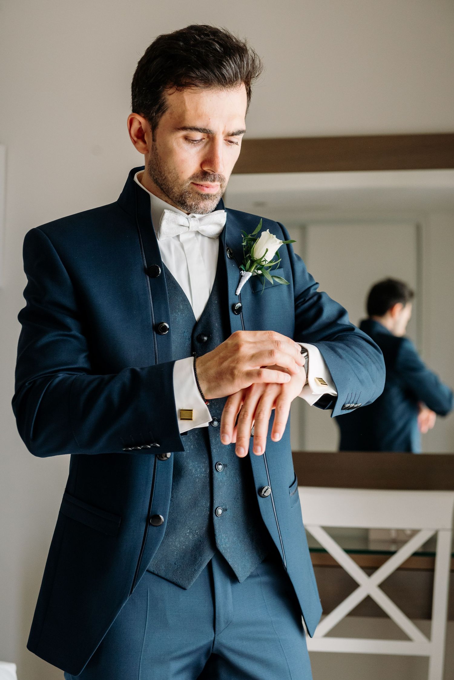 Groom in navy suit adjusts boutonniere and cuffs in elegant indoor setting.