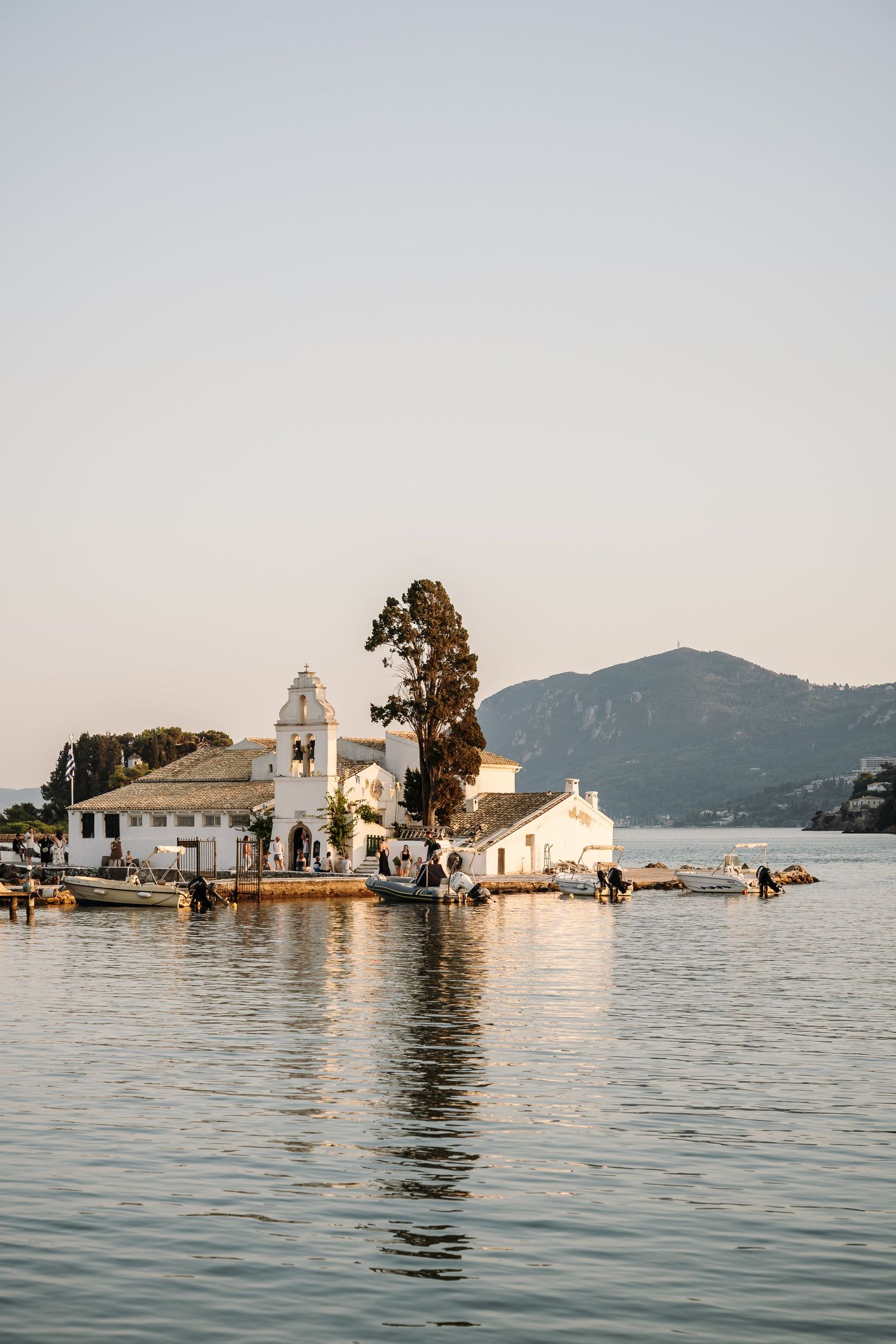 A white Greek Orthodox church with bell tower sits on the waterfront of a Mediterranean island.