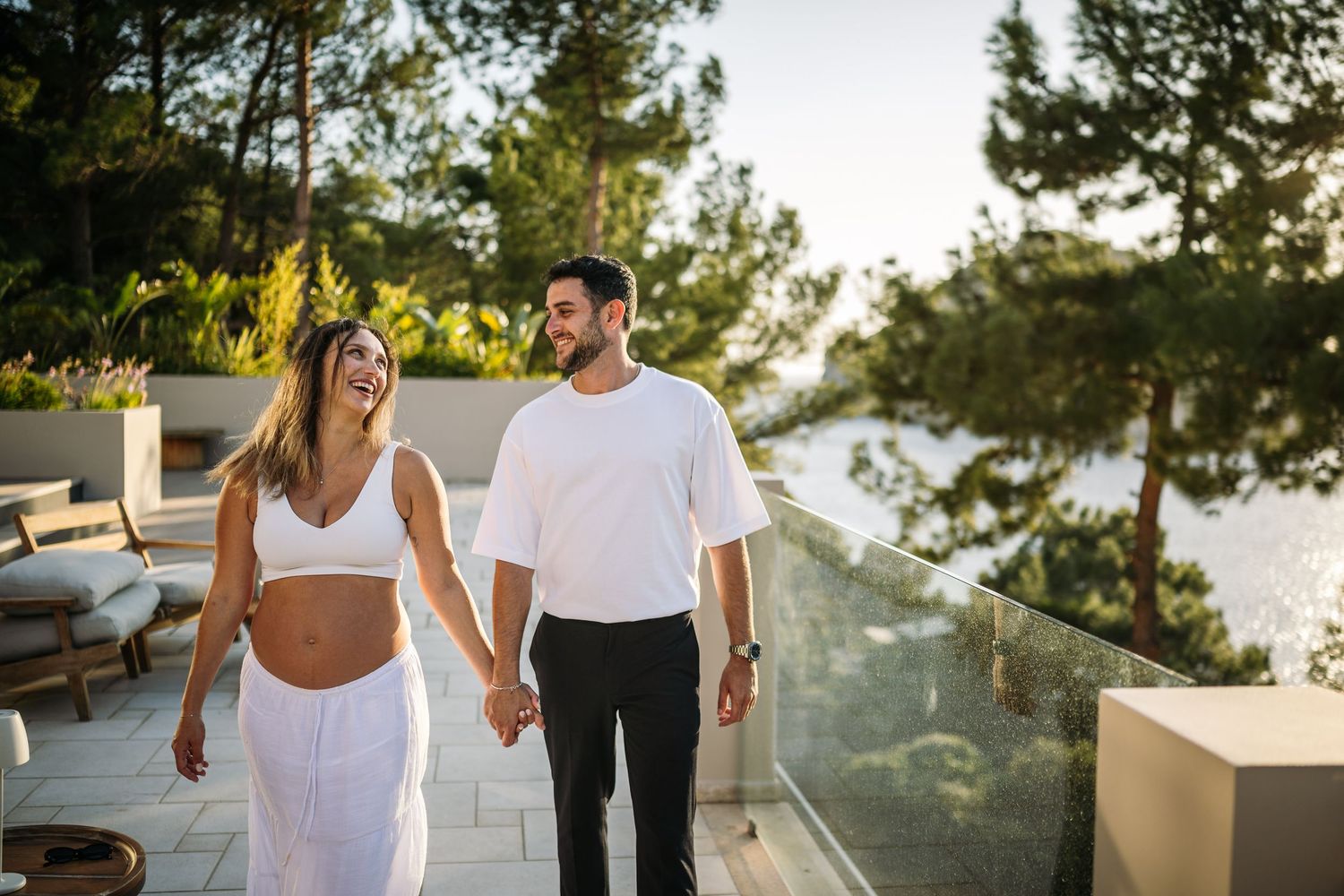 A modern couple in white outfits walking on a terrace with scenic mountain and ocean views at sunset.
