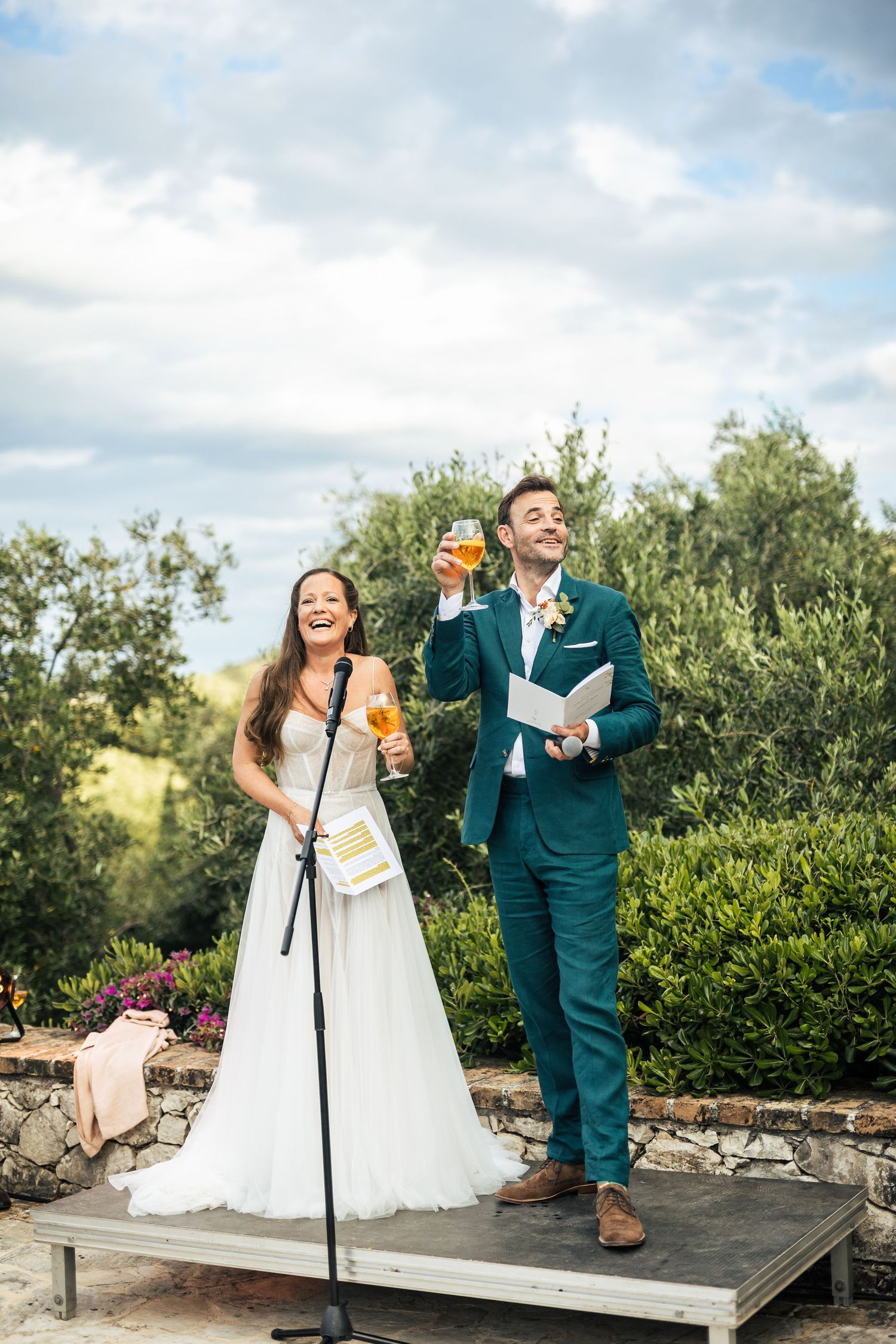 A couple shares a joyful moment while reading from a book during an outdoor wedding ceremony.