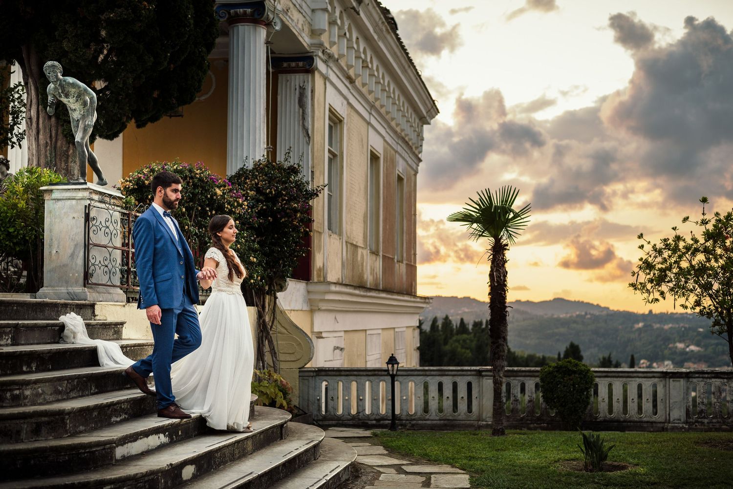 Elegant wedding couple poses against a dramatic sunset backdrop at a grand estate.