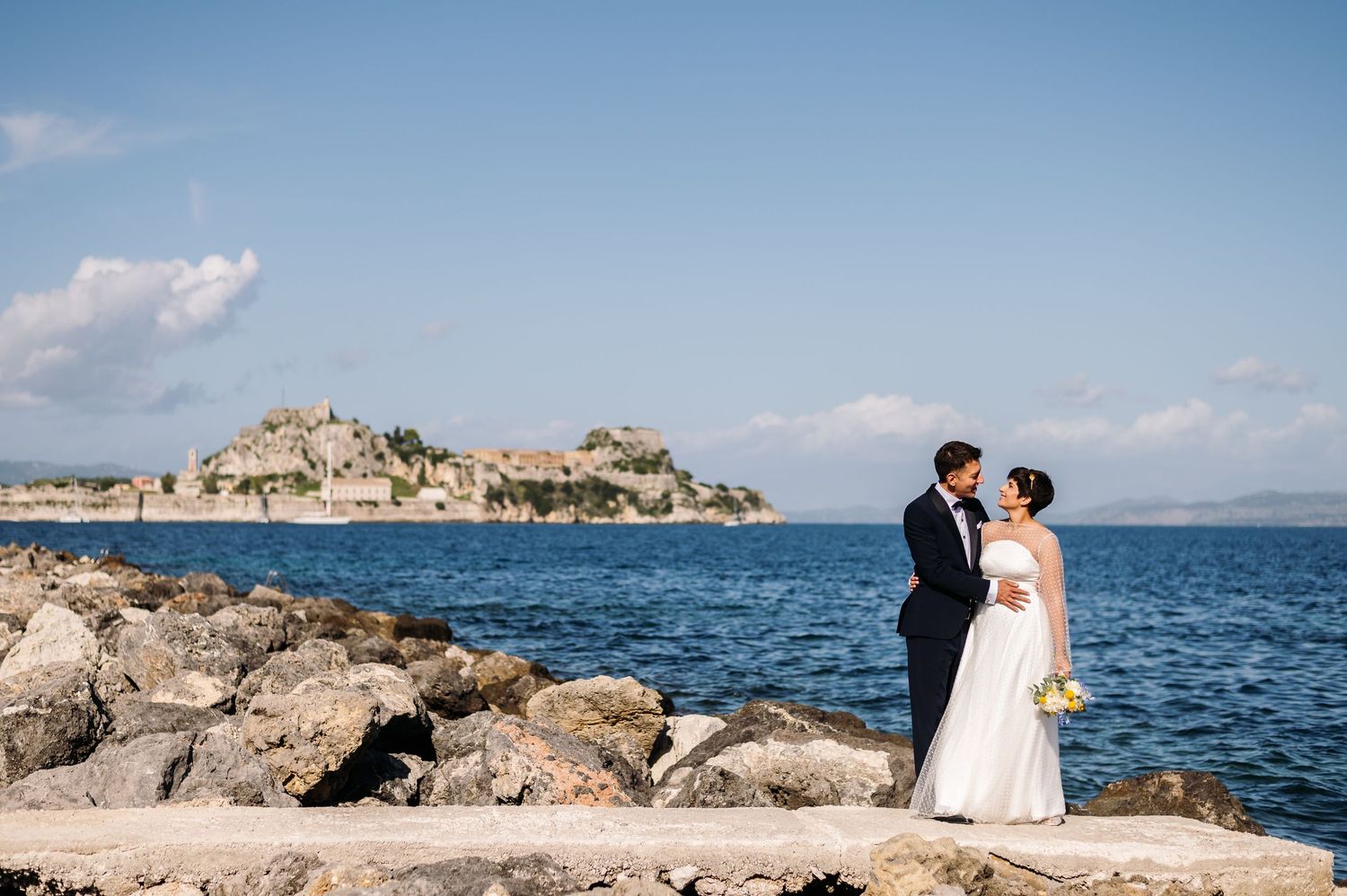 A wedding couple poses on rocky coastal cliffs overlooking the blue ocean.