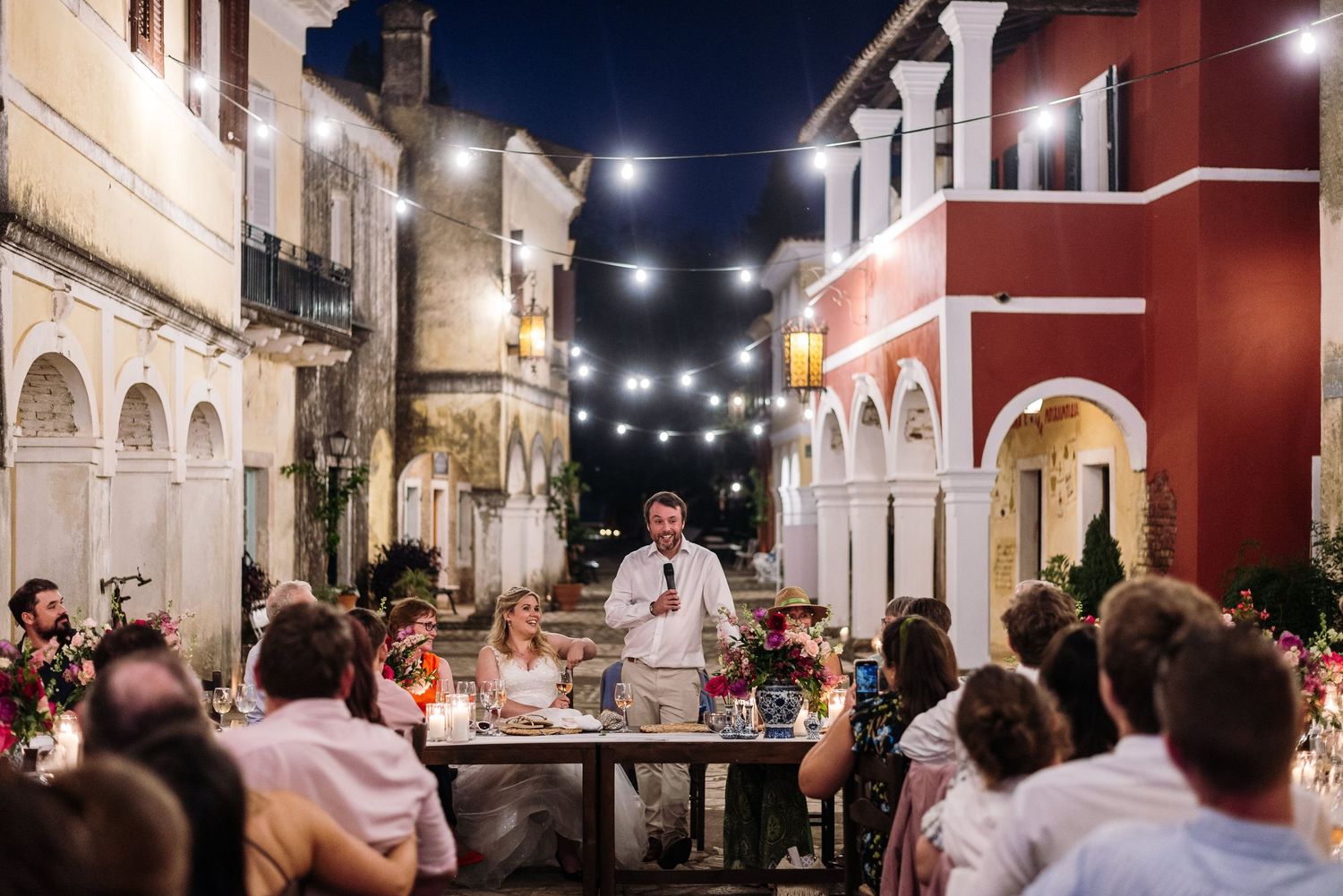 Nighttime outdoor wedding reception with string lights illuminating historic colonial architecture.