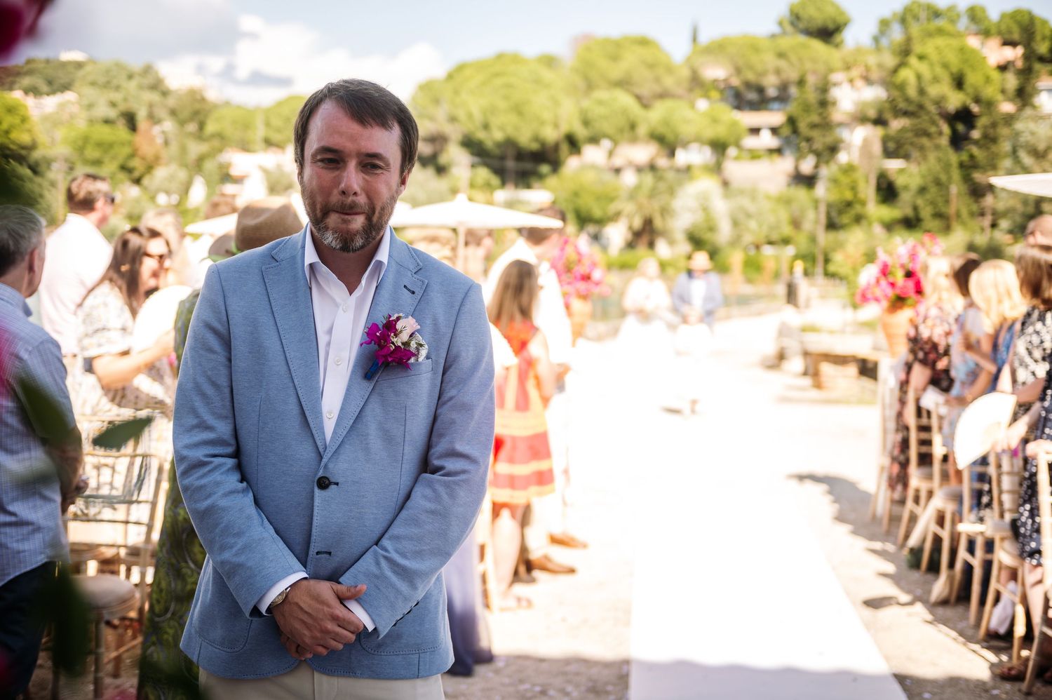 Sunny outdoor wedding ceremony with guests seated in wooden chairs.