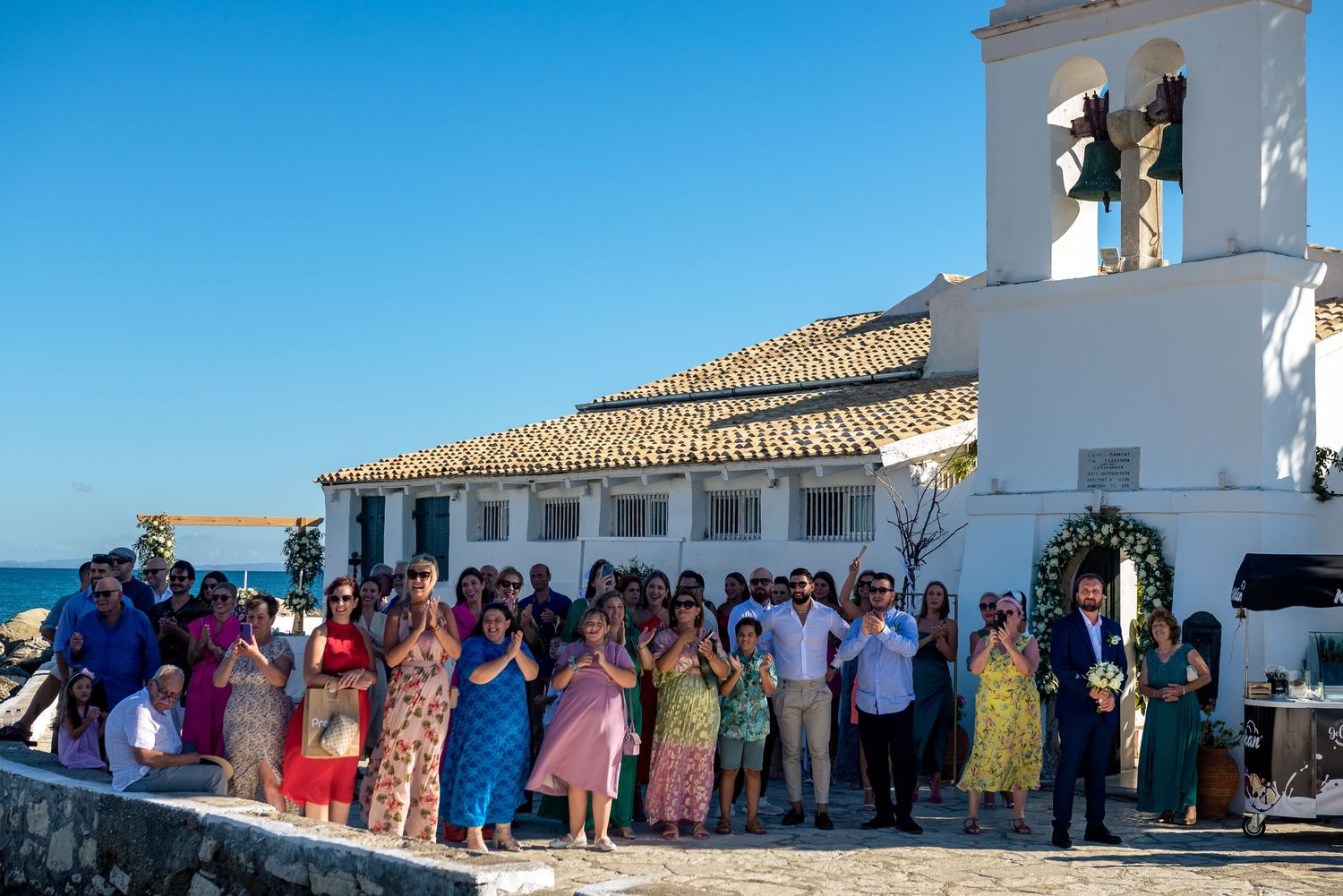 Wedding ceremony guests gather outside a white Mediterranean-style church building under blue skies.