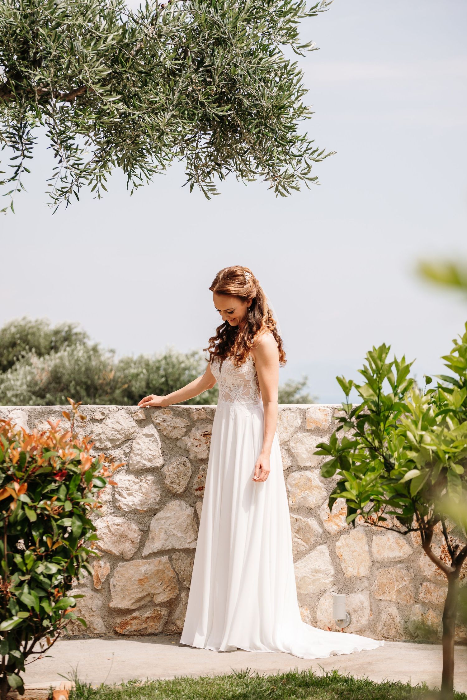 A flowing white wedding dress photographed against a rustic stone wall and olive trees in a Mediterranean setting.