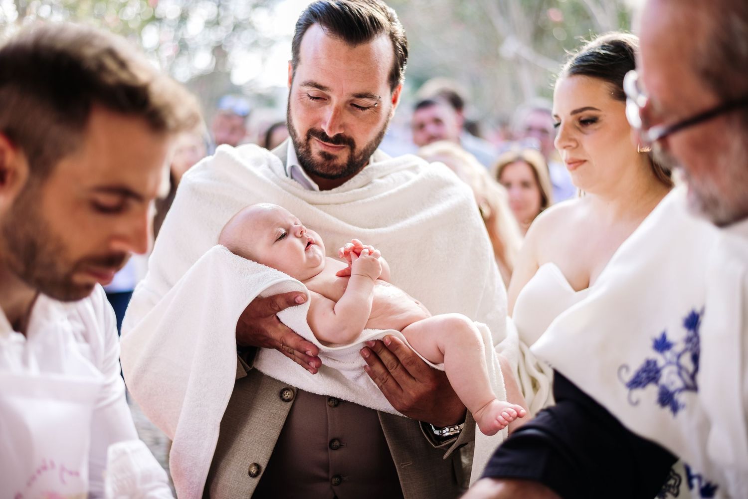 An infant being held during a baptism ceremony surrounded by family and clergy in white robes.