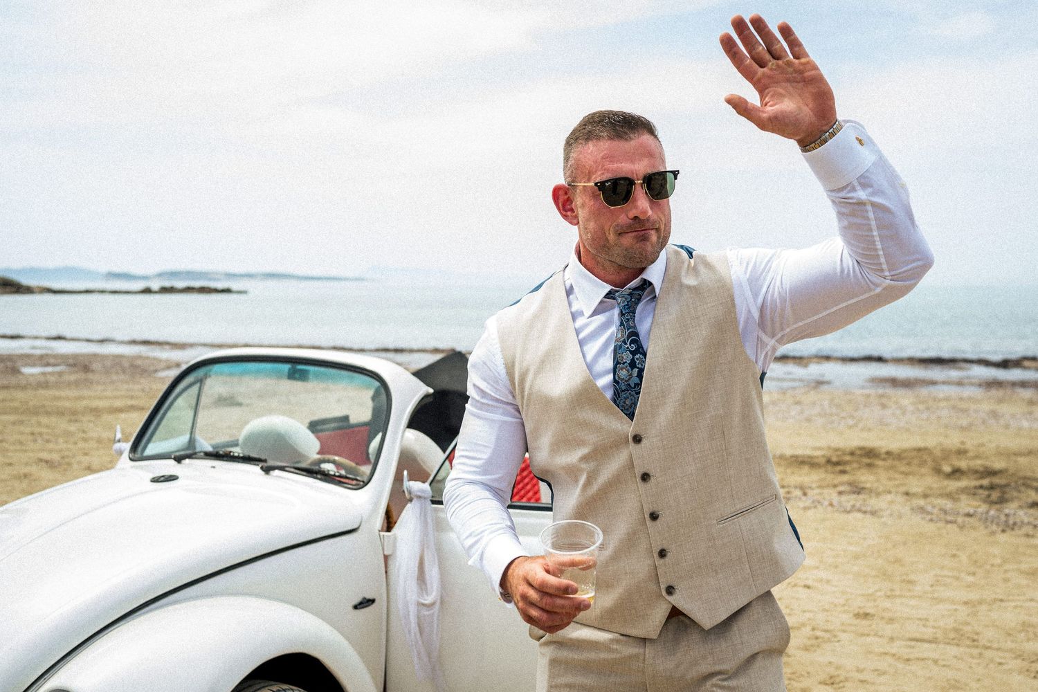 Wedding party members waving while standing near a vintage car on a beach.