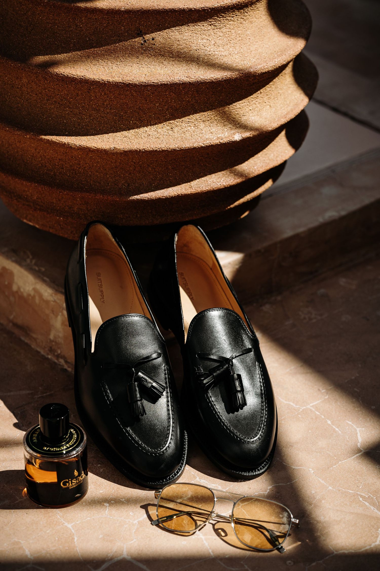 Black leather tassel loafers with sunglasses and perfume bottle in dramatic lighting.