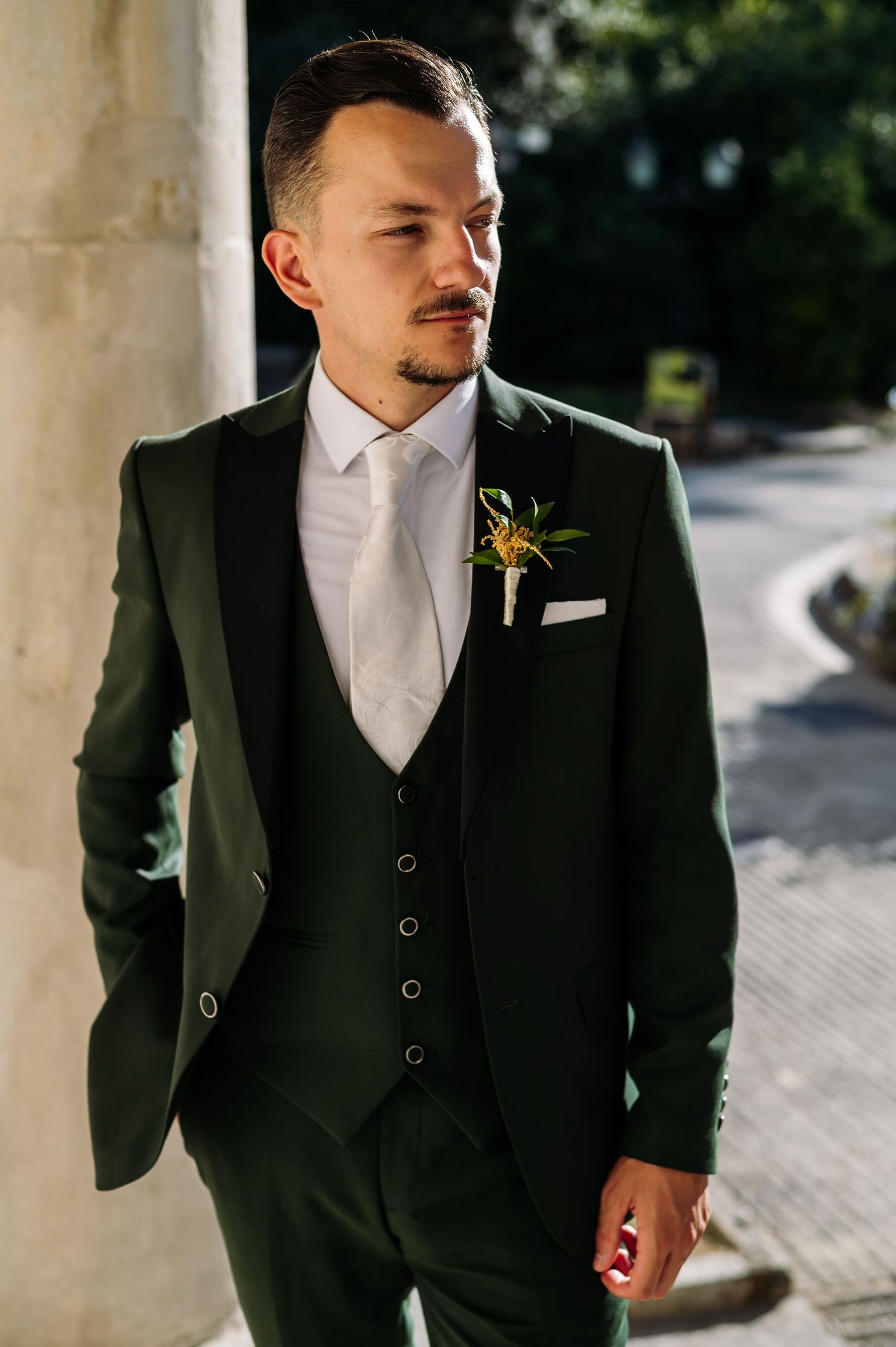 Groom in classic black three-piece suit stands in sunlit colonnade.