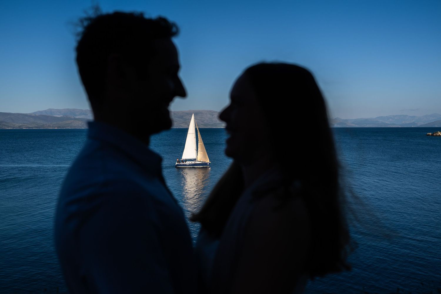 Silhouettes against a blue ocean backdrop with a small sailboat visible in the distance.