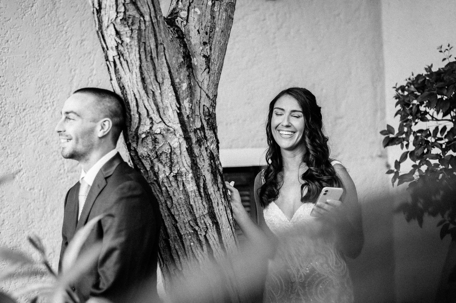 Black and white wedding photo of a couple sharing a joyful moment while standing separated by a tree.