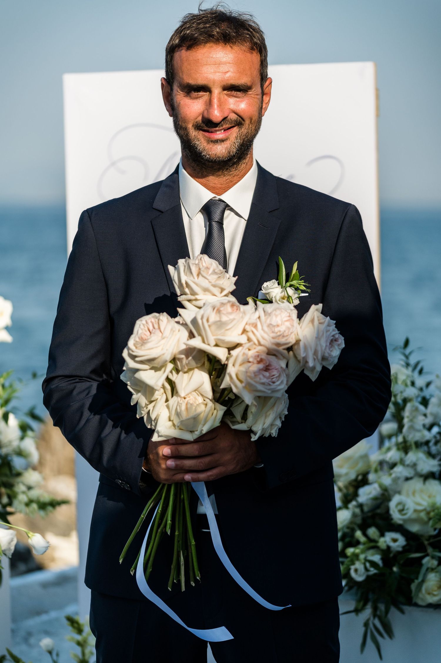 Groom in black suit holding a luxurious white rose bouquet against an ocean backdrop.