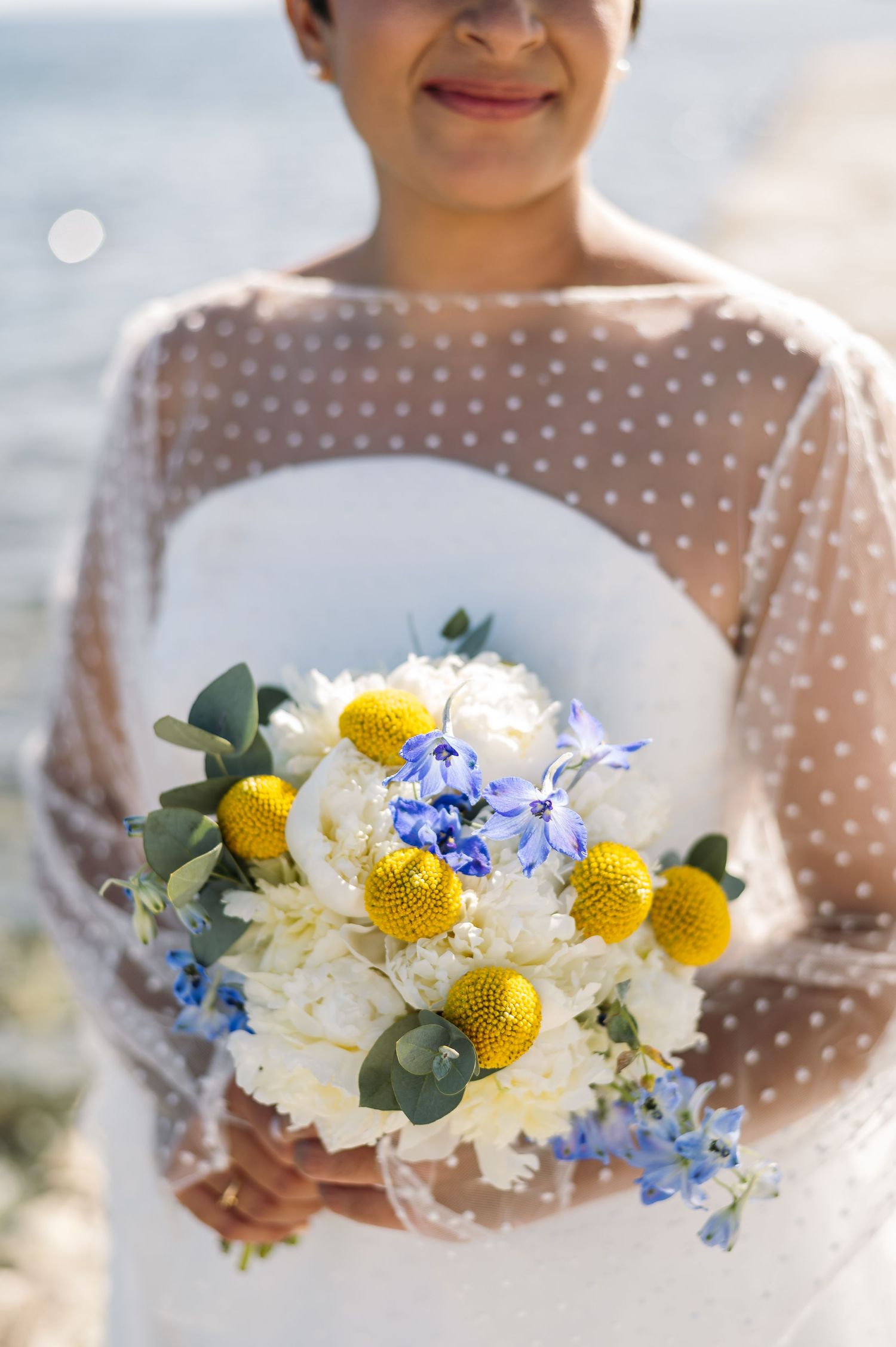 Wedding bouquet featuring yellow craspedia, white roses, and blue delphinium flowers.