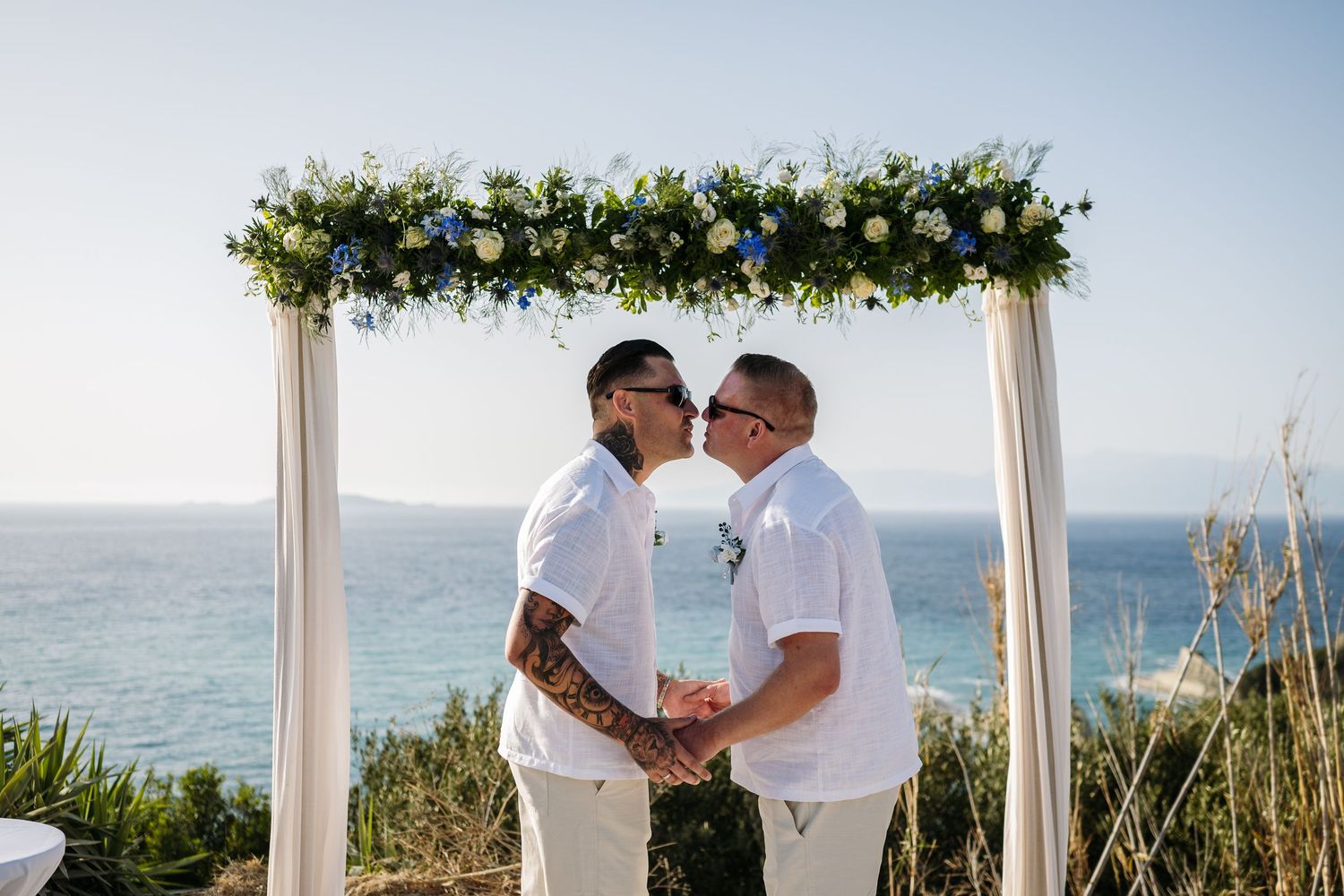 White wedding arch with greenery stands against blue ocean backdrop during beach ceremony.
