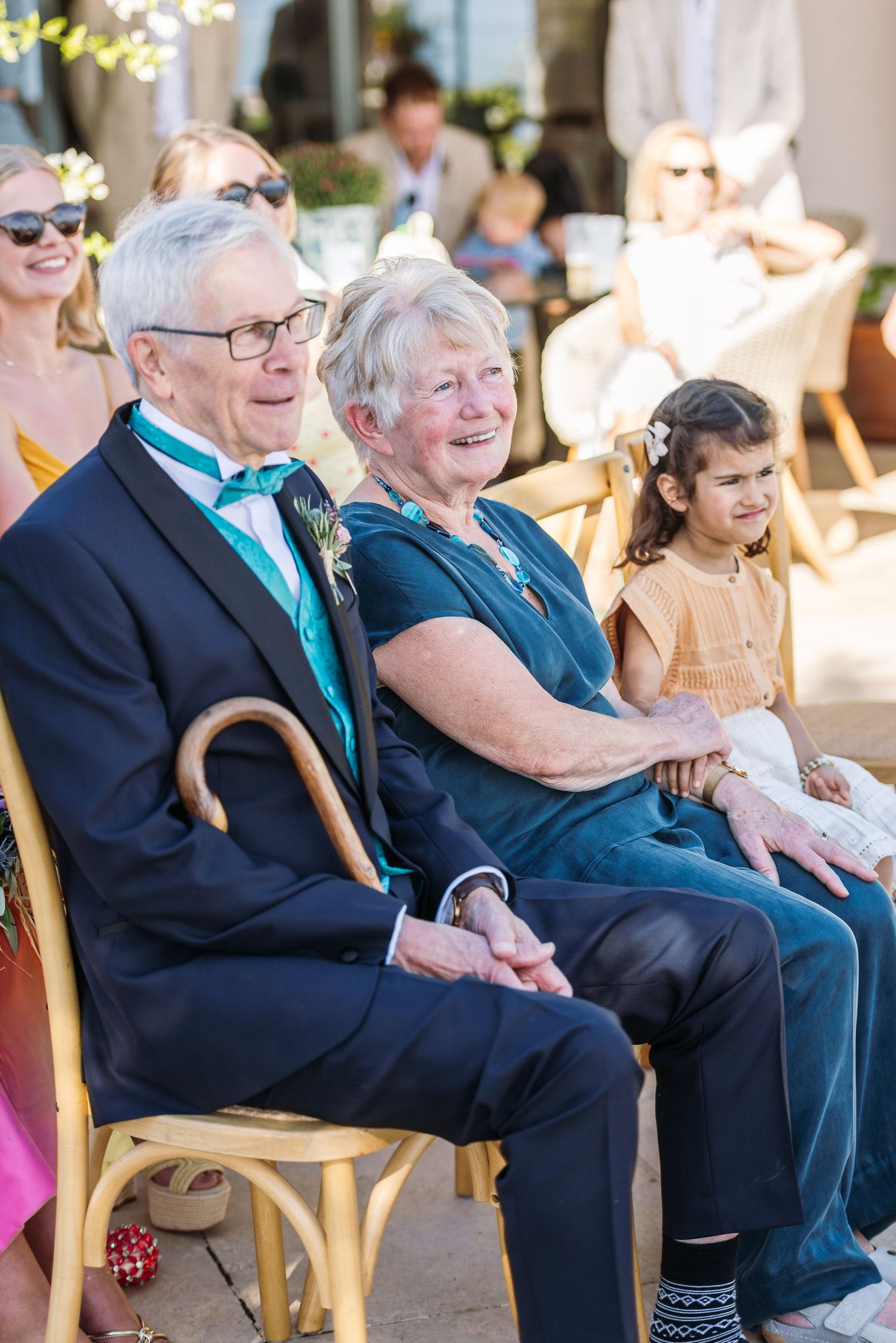 Wedding guests seated in wooden chairs smile and enjoy an outdoor ceremony.