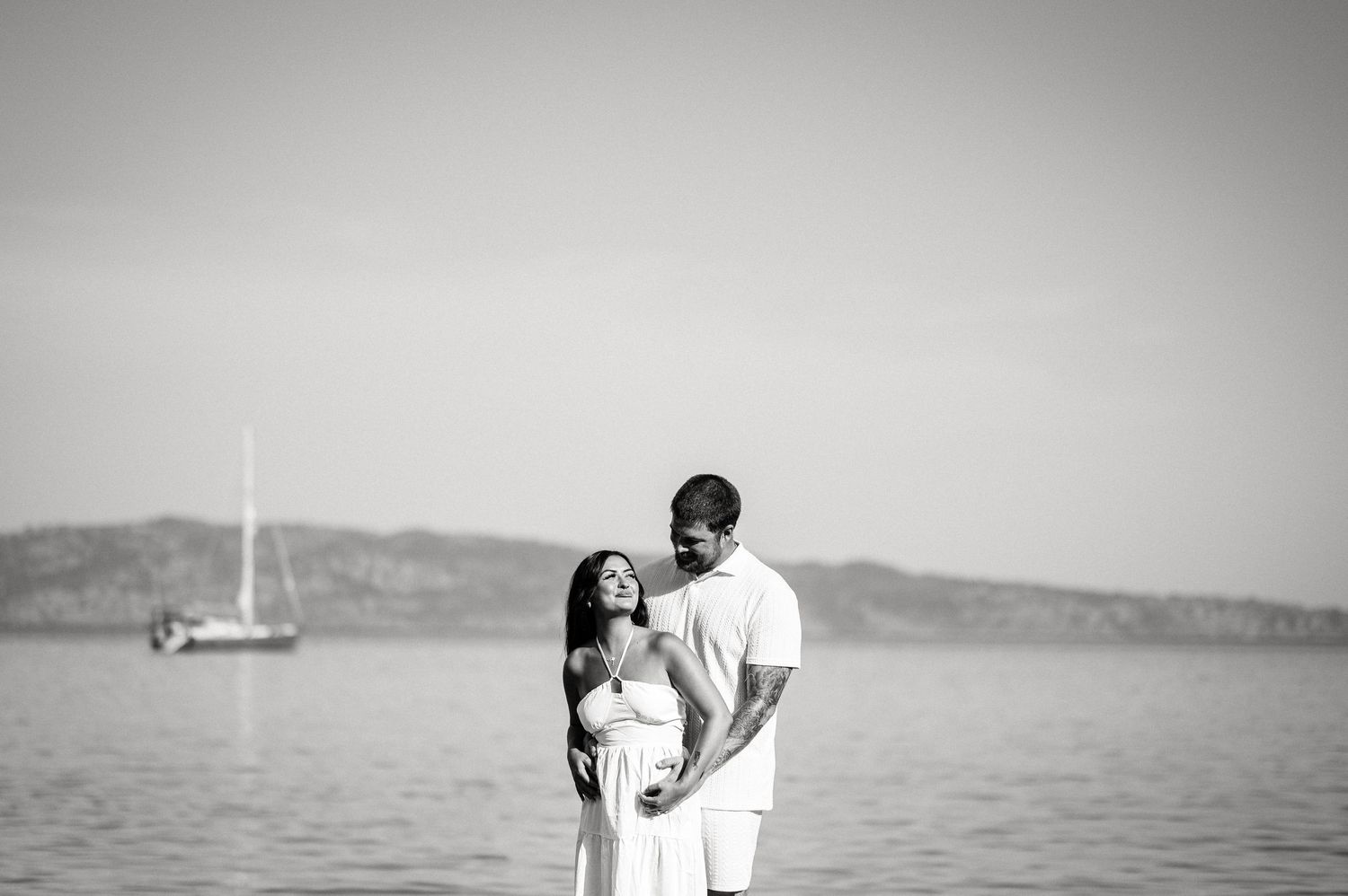 A black and white portrait of a couple in white clothing standing by the sea with a sailboat in the distance.