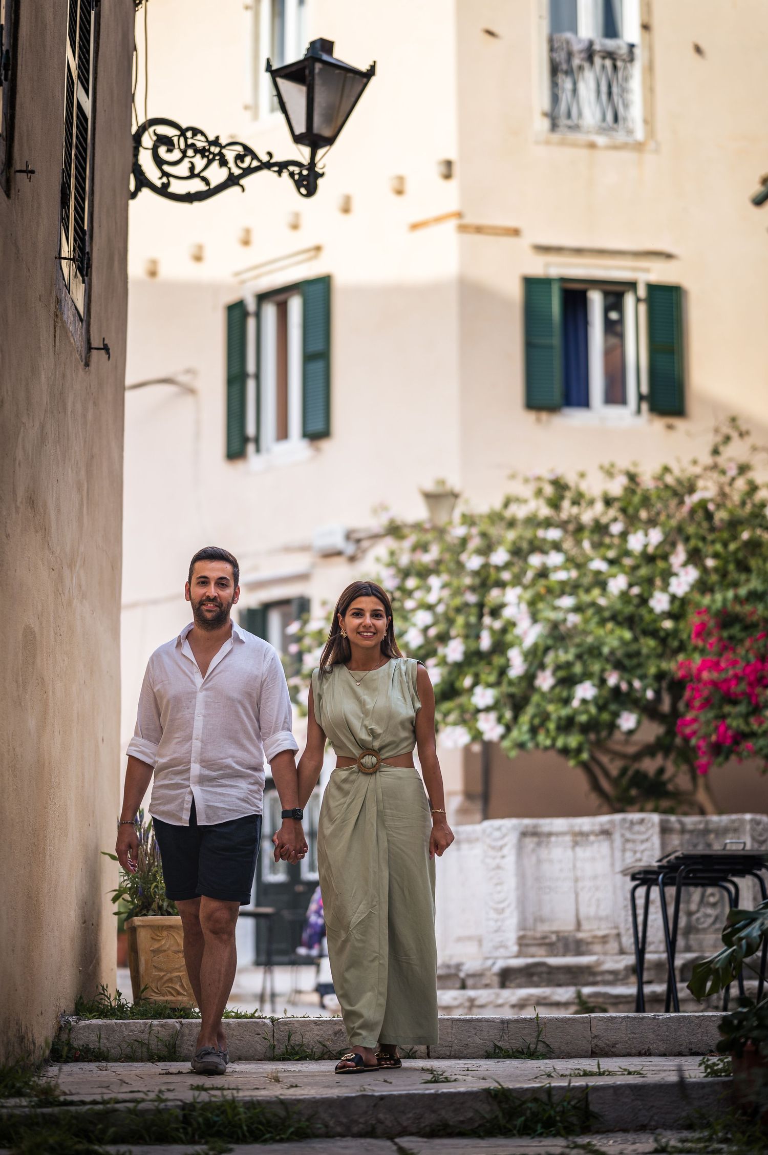 A couple strolls down charming Mediterranean steps with green shuttered buildings and flowering bushes in the background.