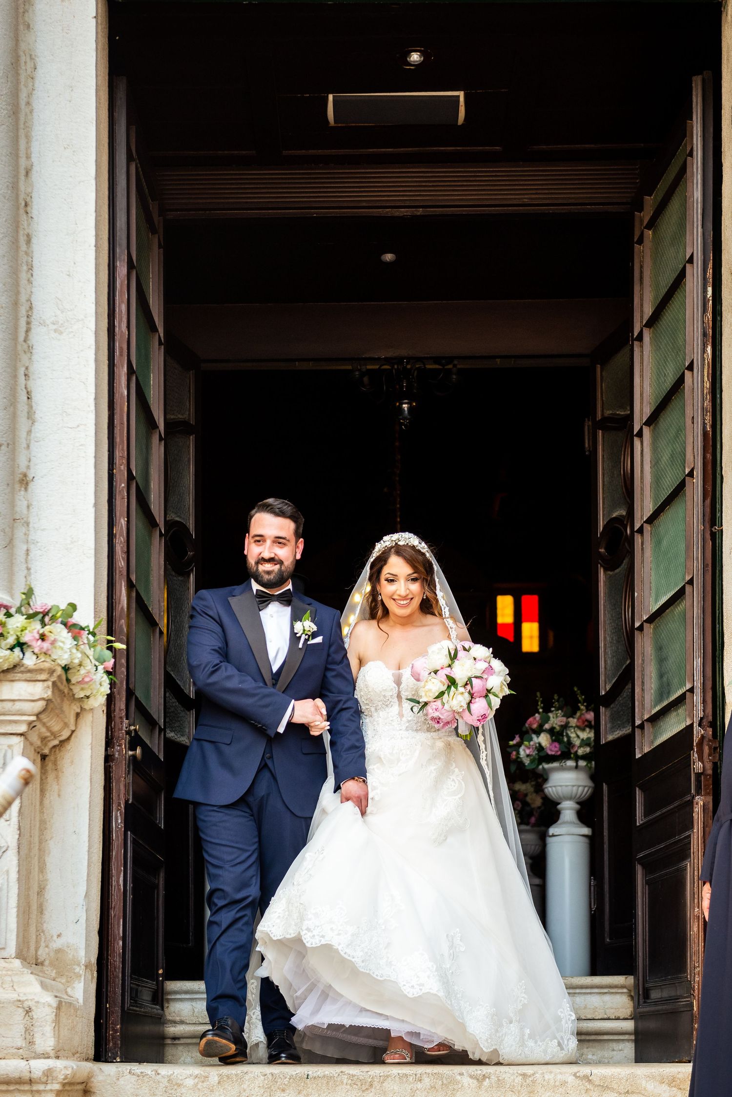 A wedding couple exits a historic building with elegant white columns and architectural details.