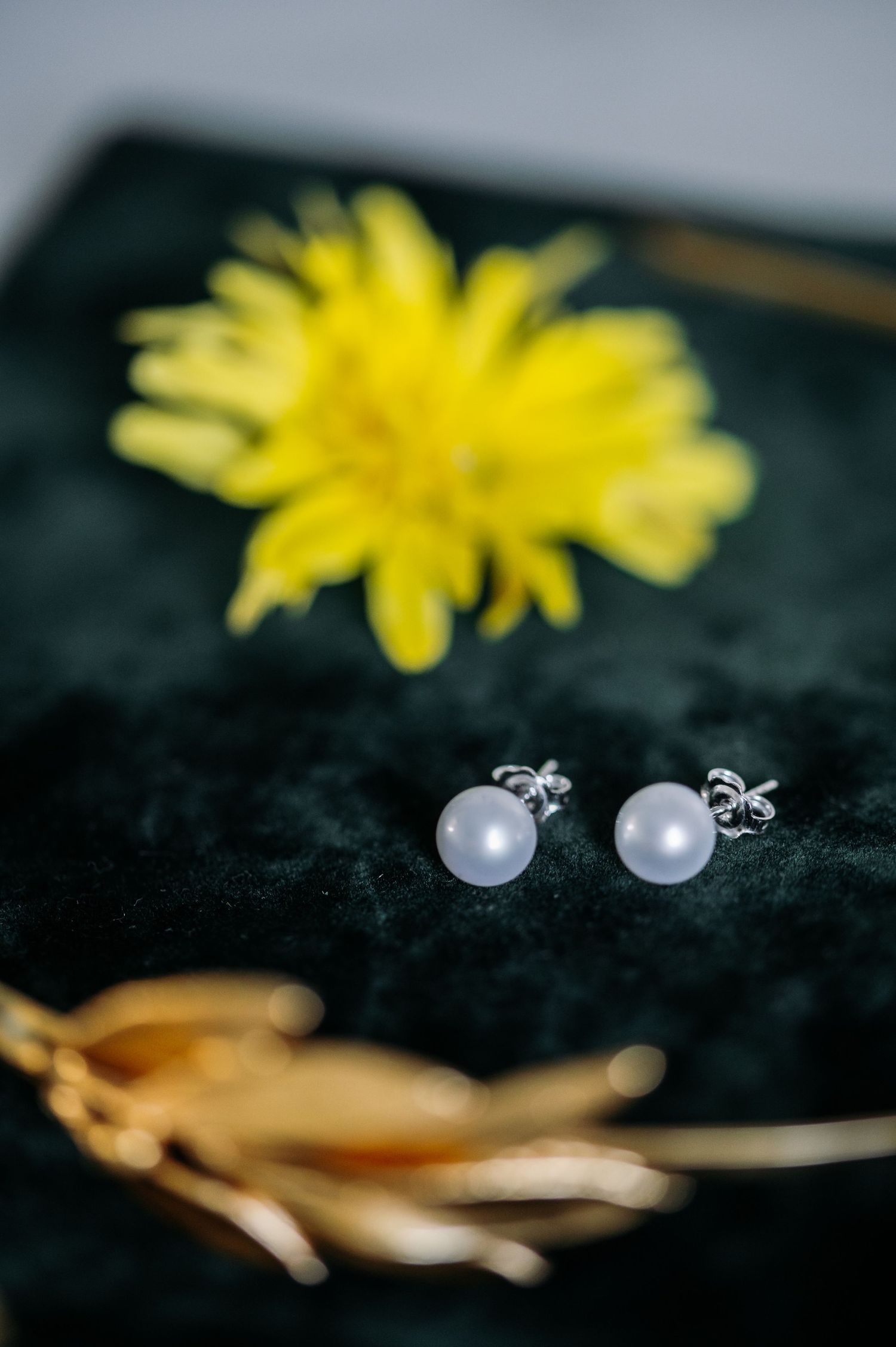 Pearl earrings and golden accessories arranged with yellow flowers on black velvet surface.
