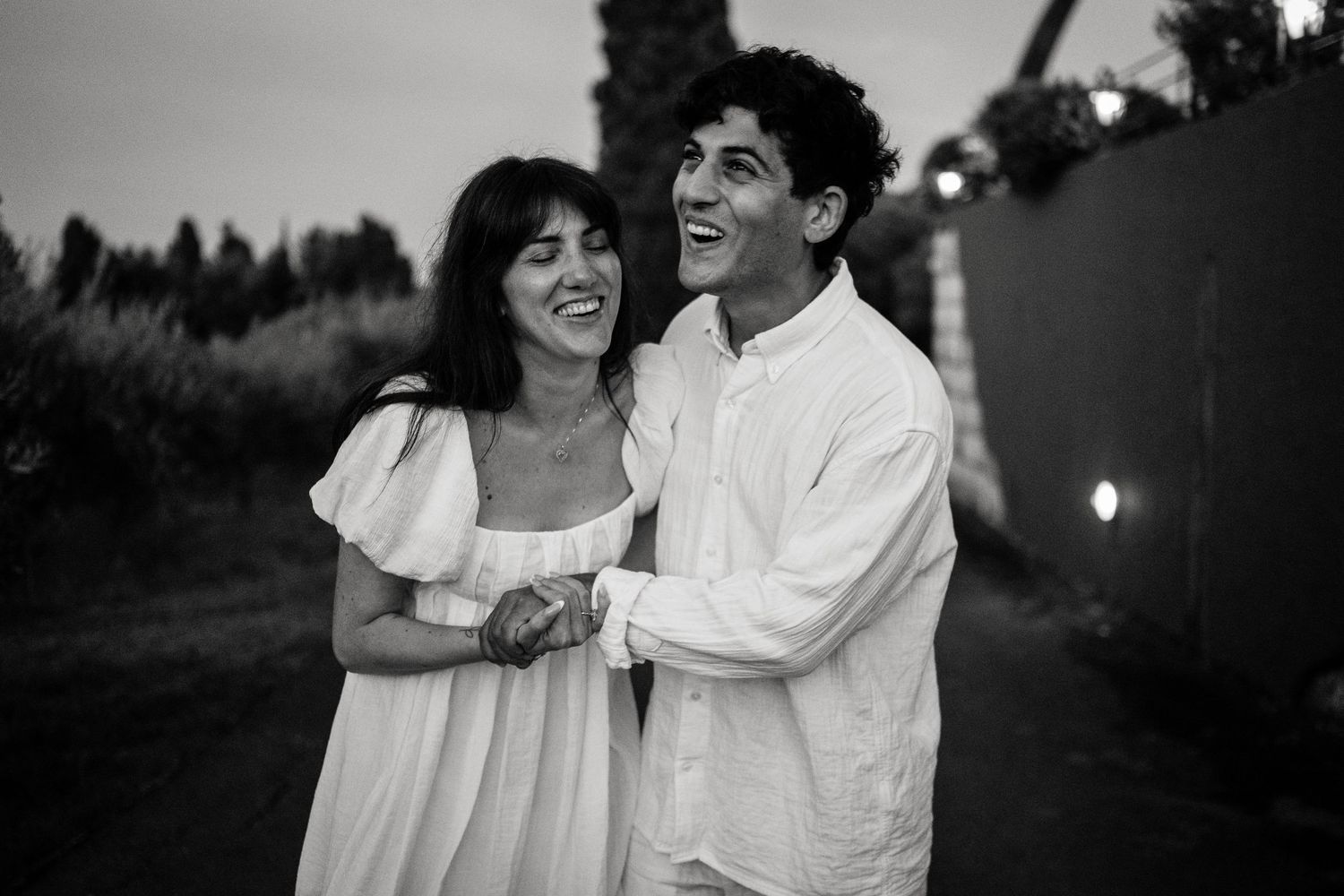 A joyful black and white photo of a couple in white clothing sharing a laugh on an evening path.