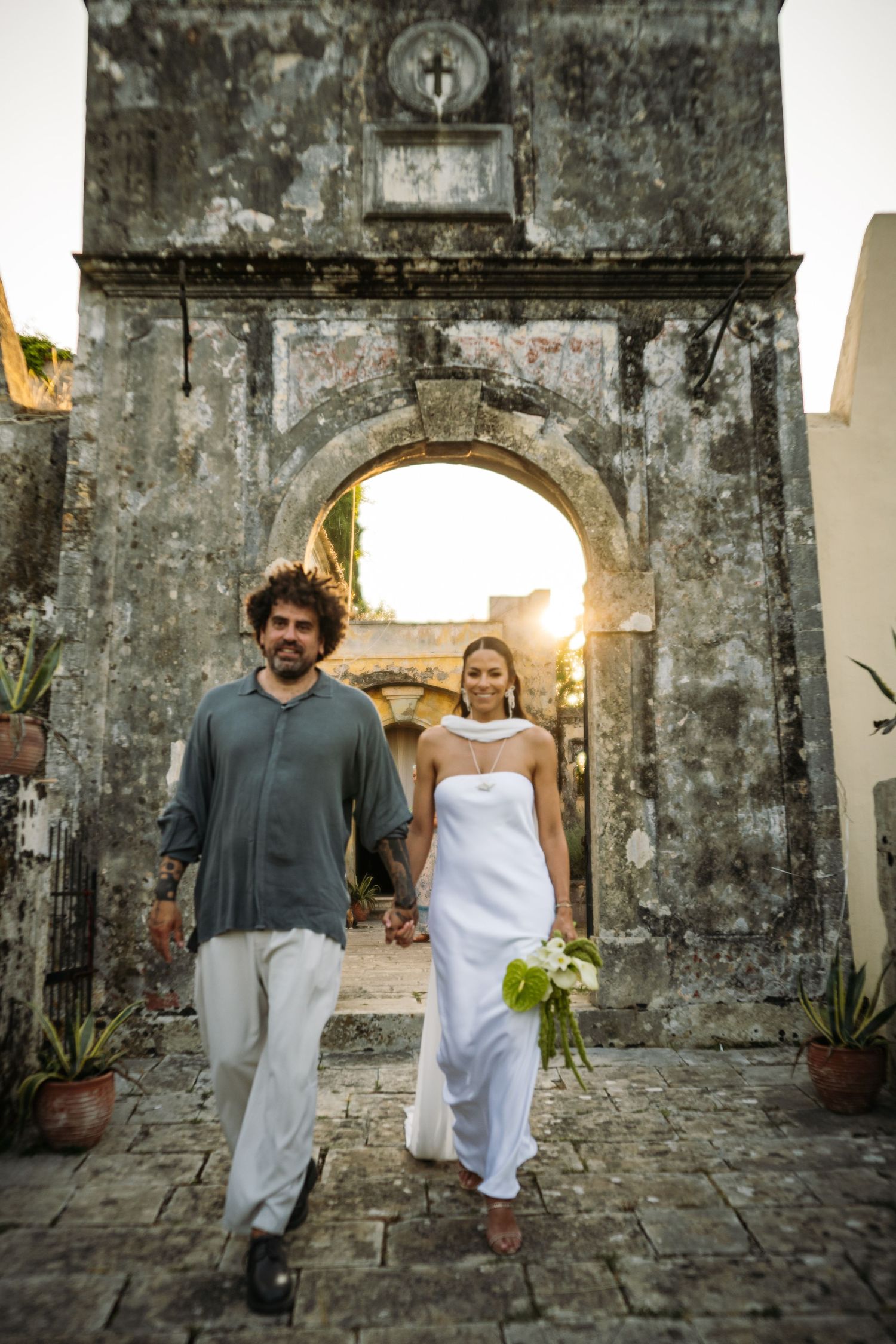 Two figures pose against a weathered stone wall with arched doorways in casual attire.