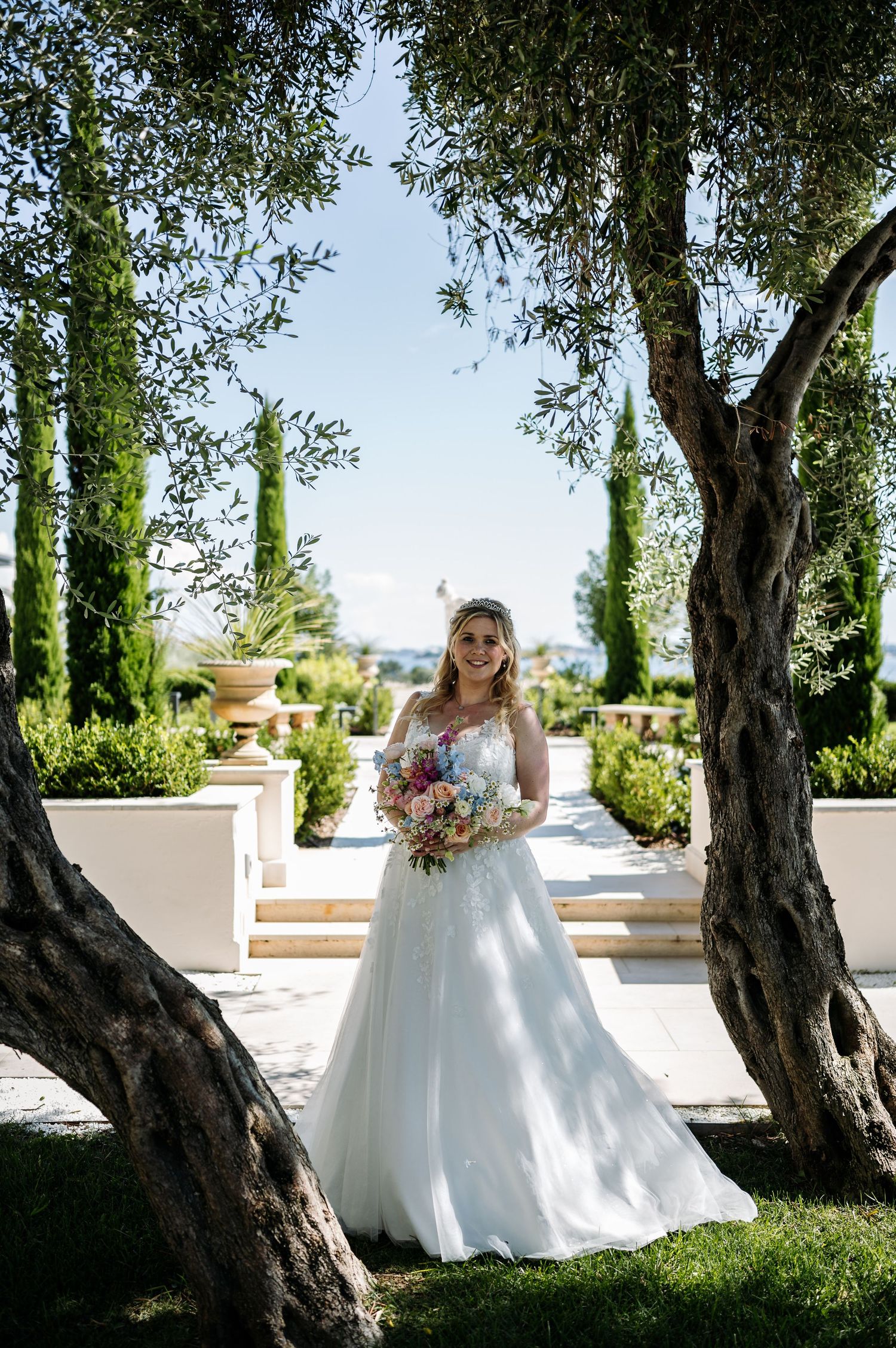 Bride in flowing white gown poses among olive trees in Mediterranean garden setting.