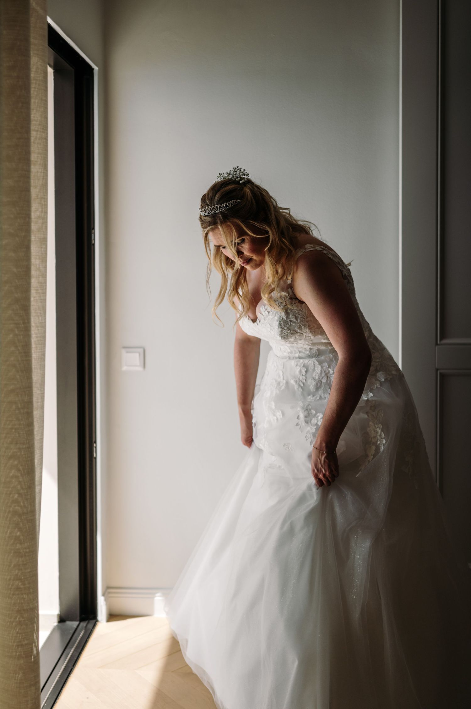 Bride in lace wedding gown standing in natural window light.