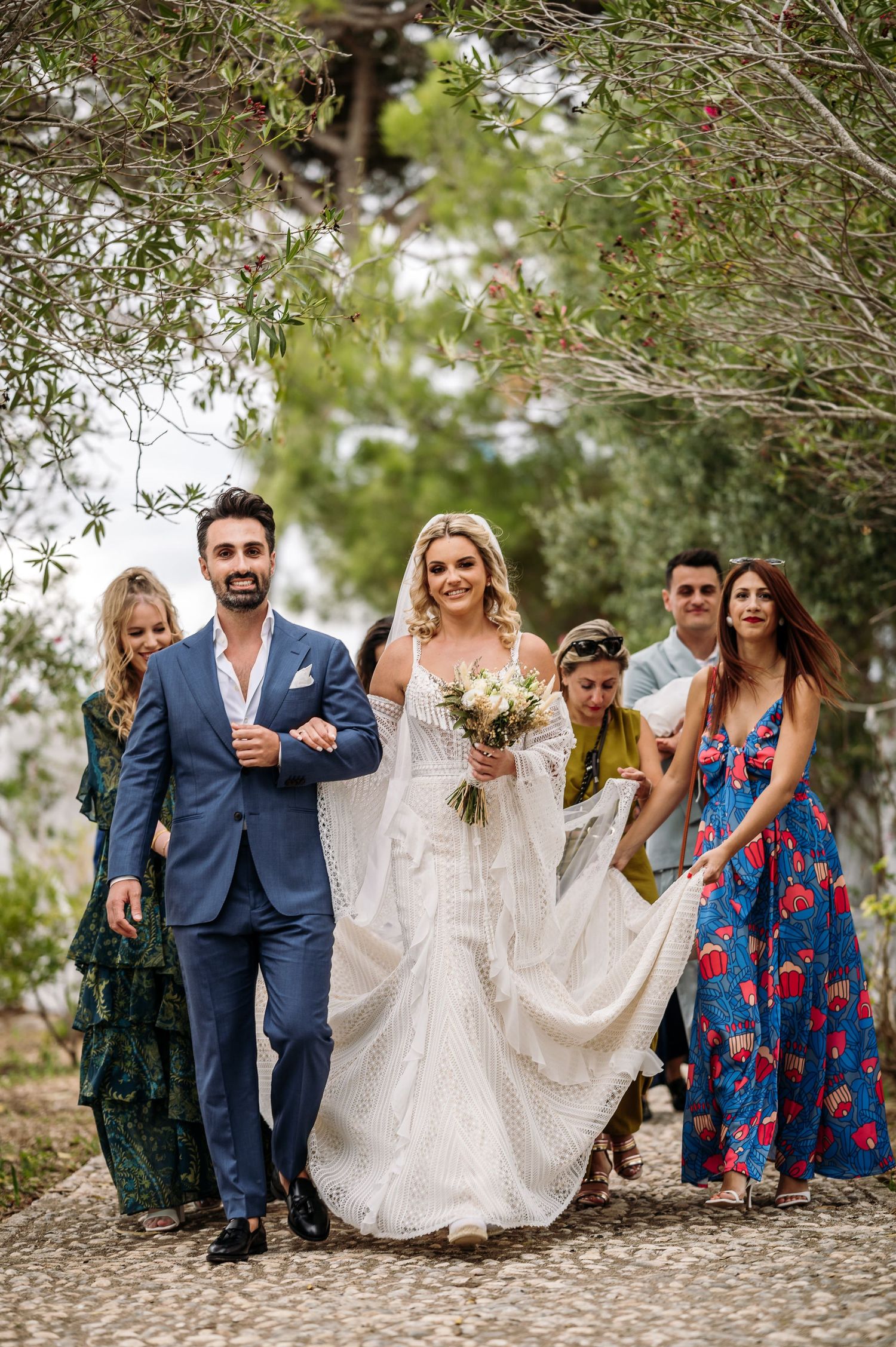 Wedding party poses for photos under trees on cobblestone path.